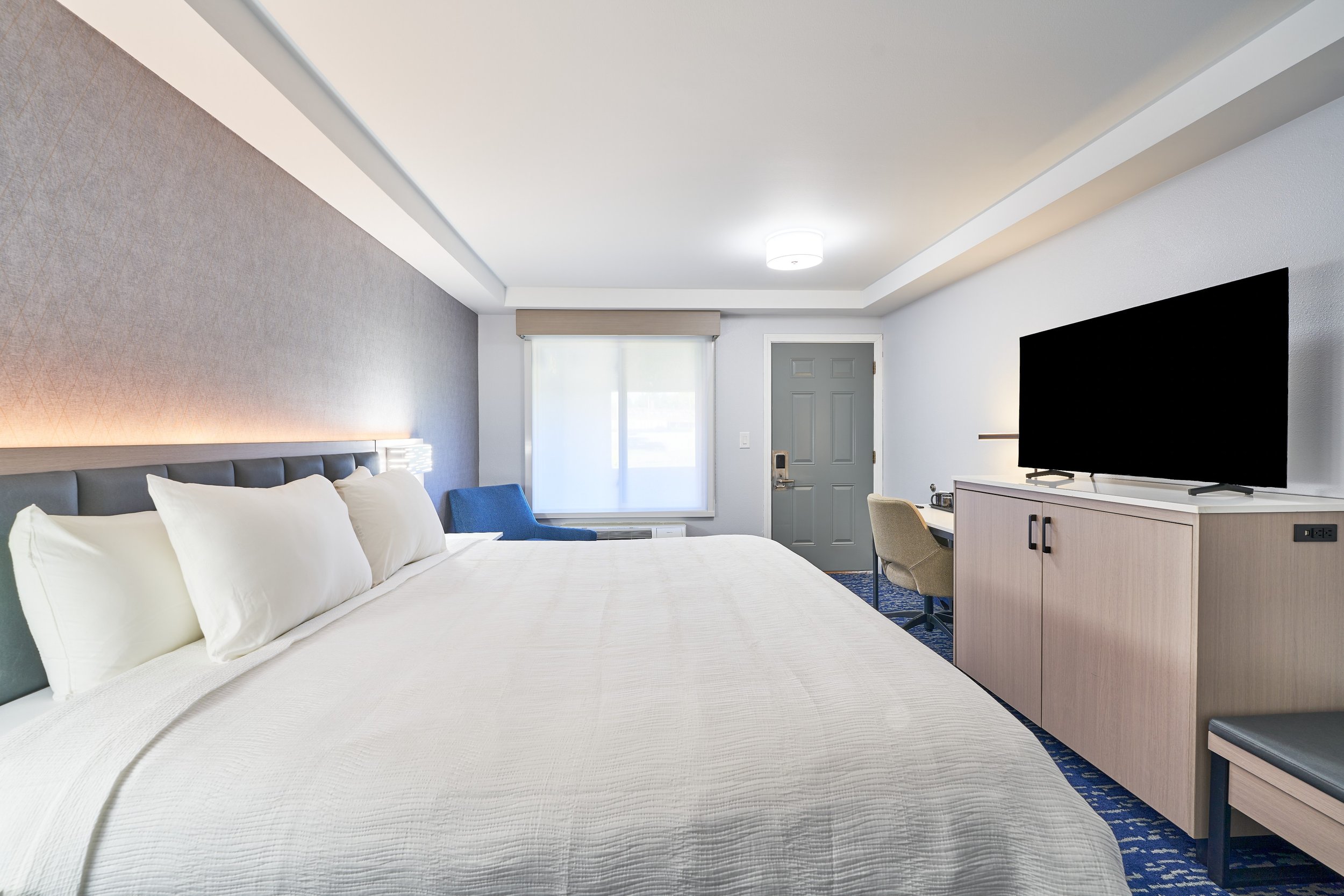 Hotel room with a large bed, a window, a grey door, a desk with a beige chair, and a flat-screen TV on a cabinet.