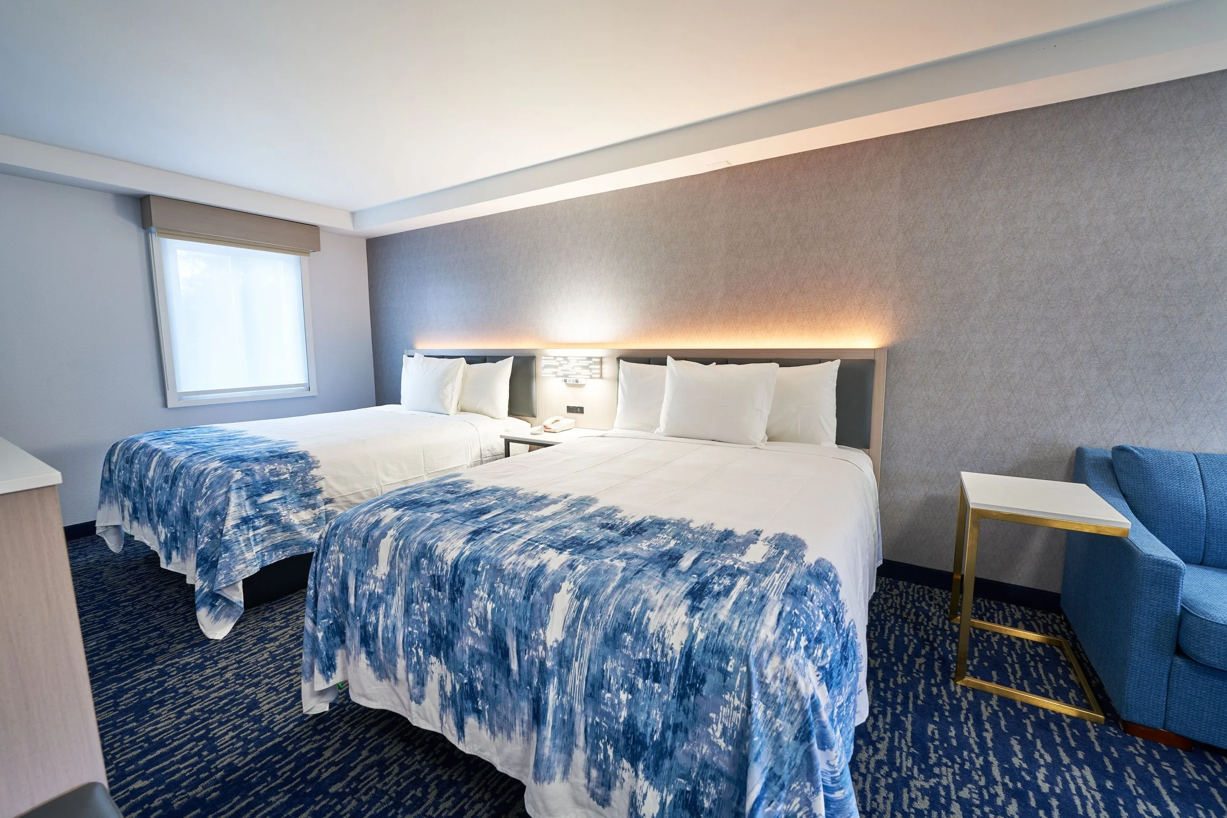 Two queen-sized beds in a hotel room with white linens, blue patterned bedspreads, a window with a beige shade, a bedside table with a telephone, a blue armchair, and a small white and gold side table.