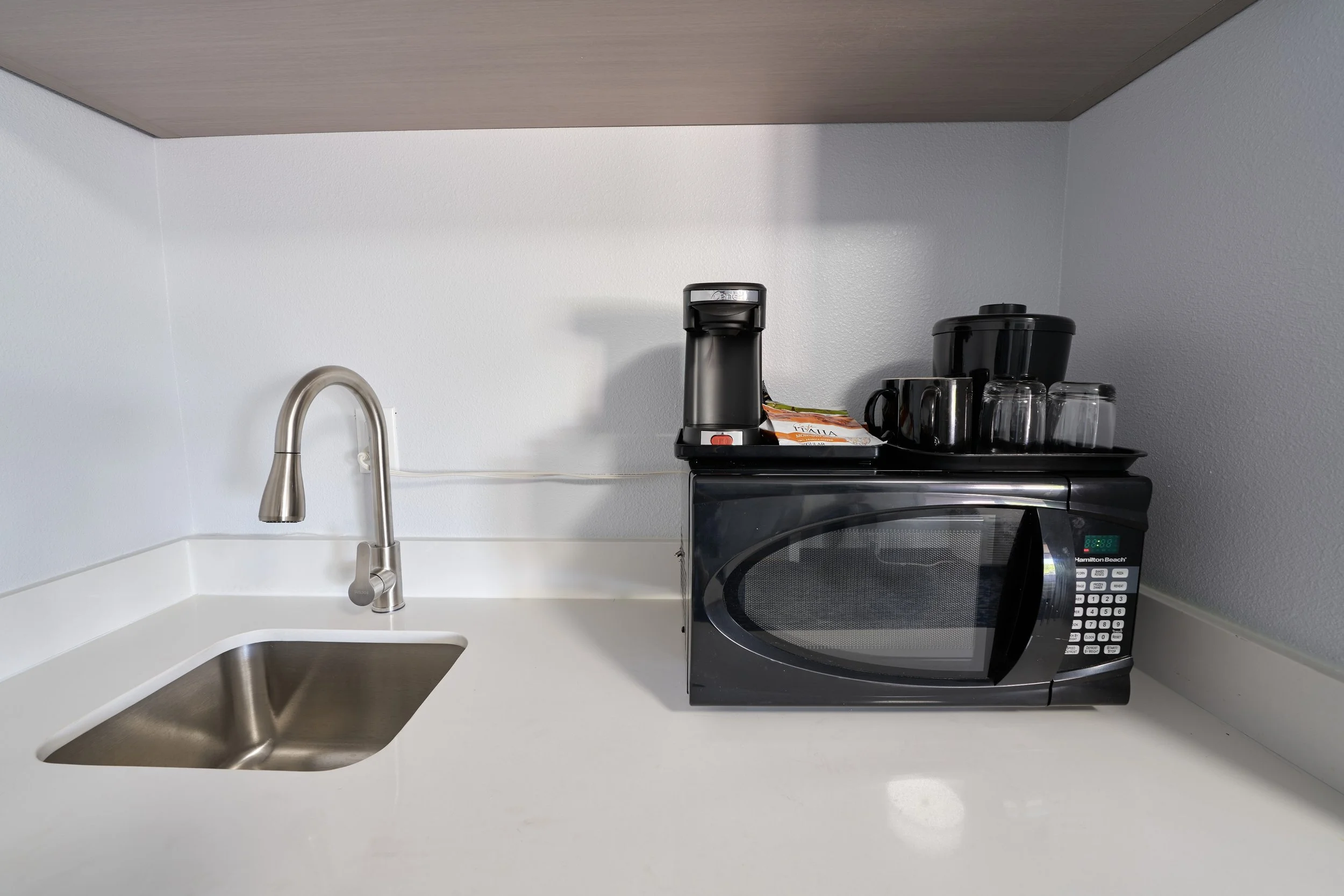 Kitchen countertop with a stainless steel sink, a silver faucet, a black microwave, a coffee maker, a black rice cooker, several cups, and a tray with packets on a white wall background.
