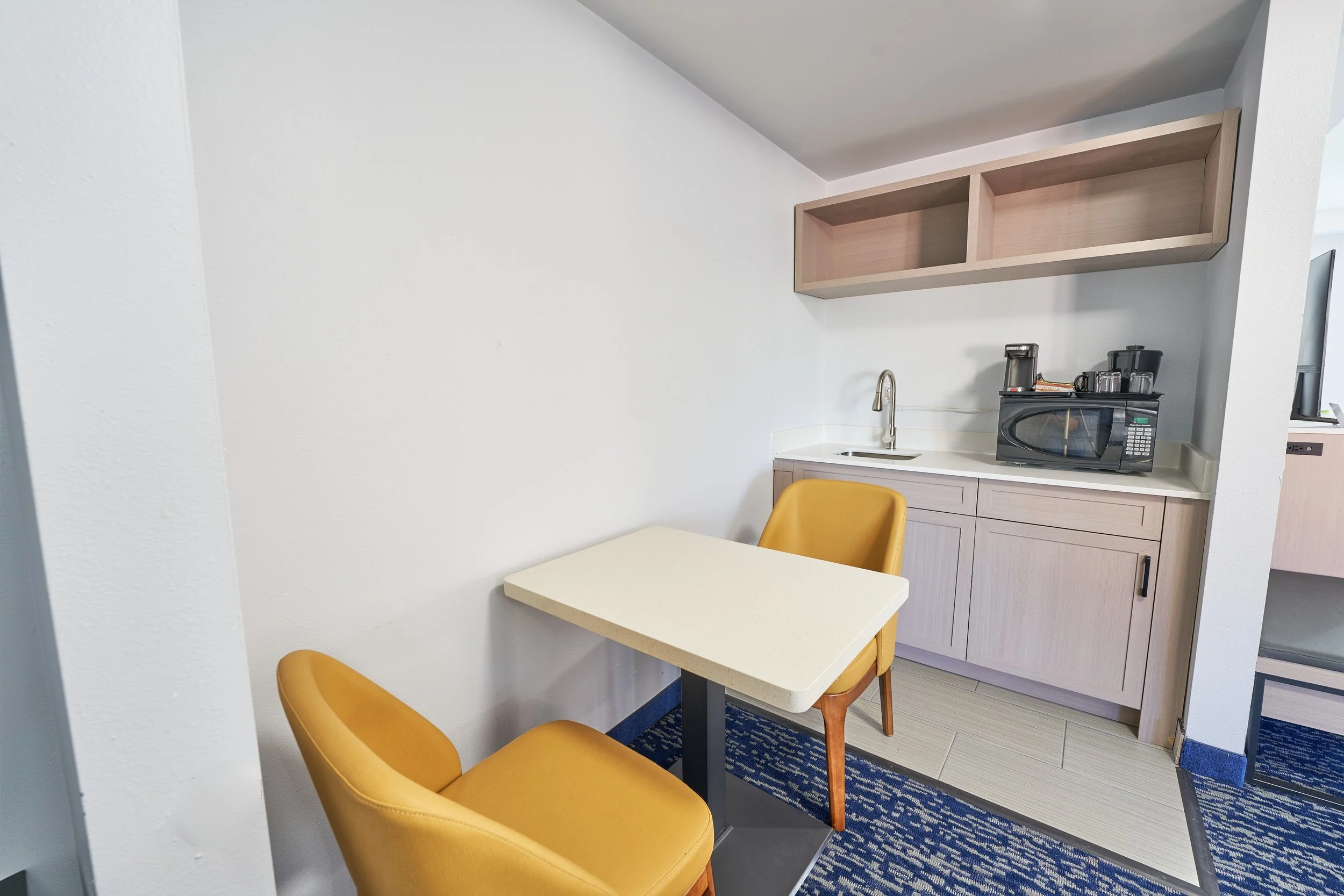 Small kitchenette with a white countertop, a sink, a microwave, coffee maker, and two open shelves above, next to a small table with two mustard-colored chairs, on a blue patterned rug.