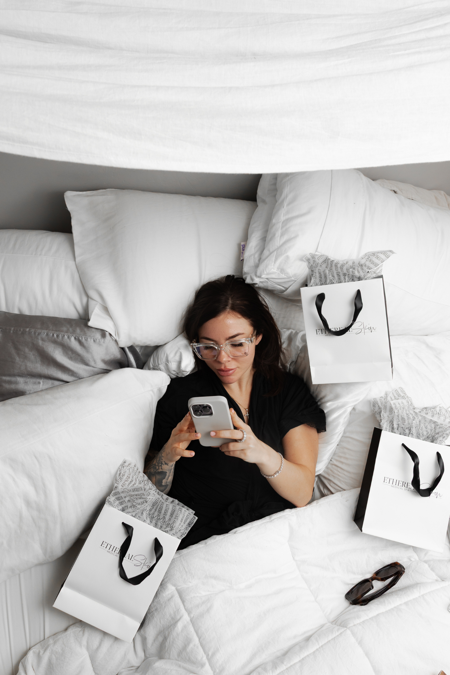 Woman on a bed scrolling her phone surrounded by shopping bags