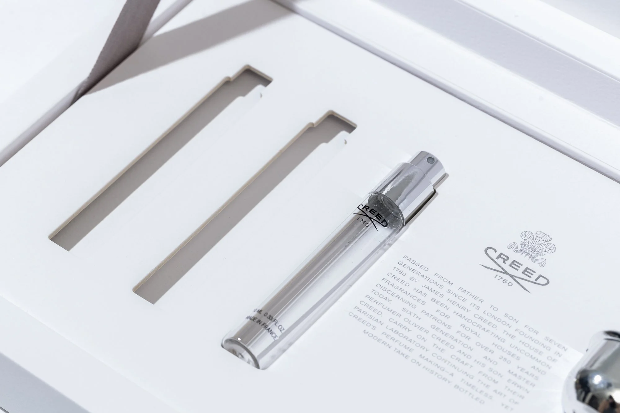 A clear glass perfume bottle with a silver cap placed on white packaging with gray text, inside a white box with cutouts.