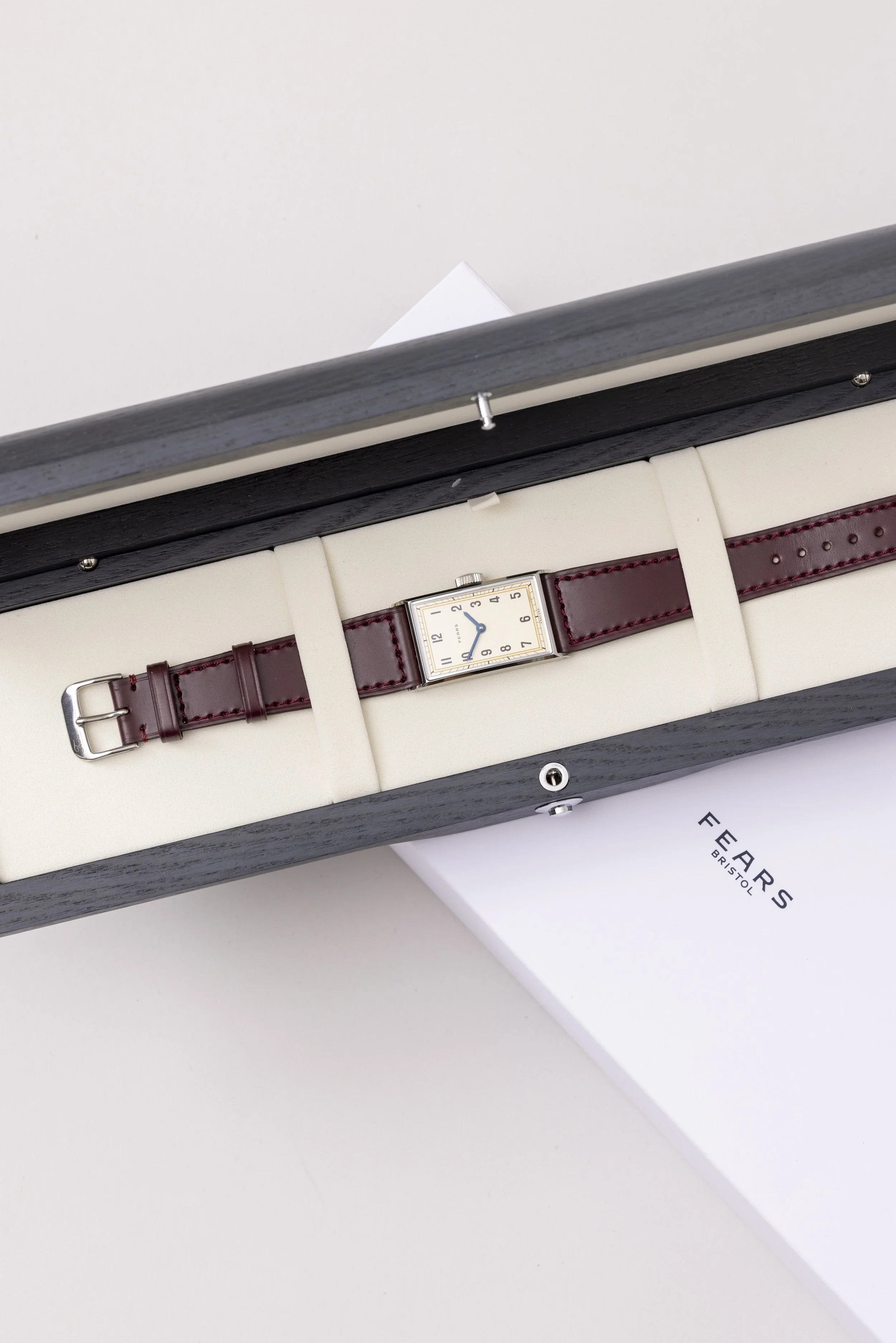 A rectangular wristwatch with a white face and brown leather strap inside a black wooden box with a cream interior. The box is partially open on a white surface, and a white rectangular card with the text 'FEARS BRISTOL' is partially visible underneath.