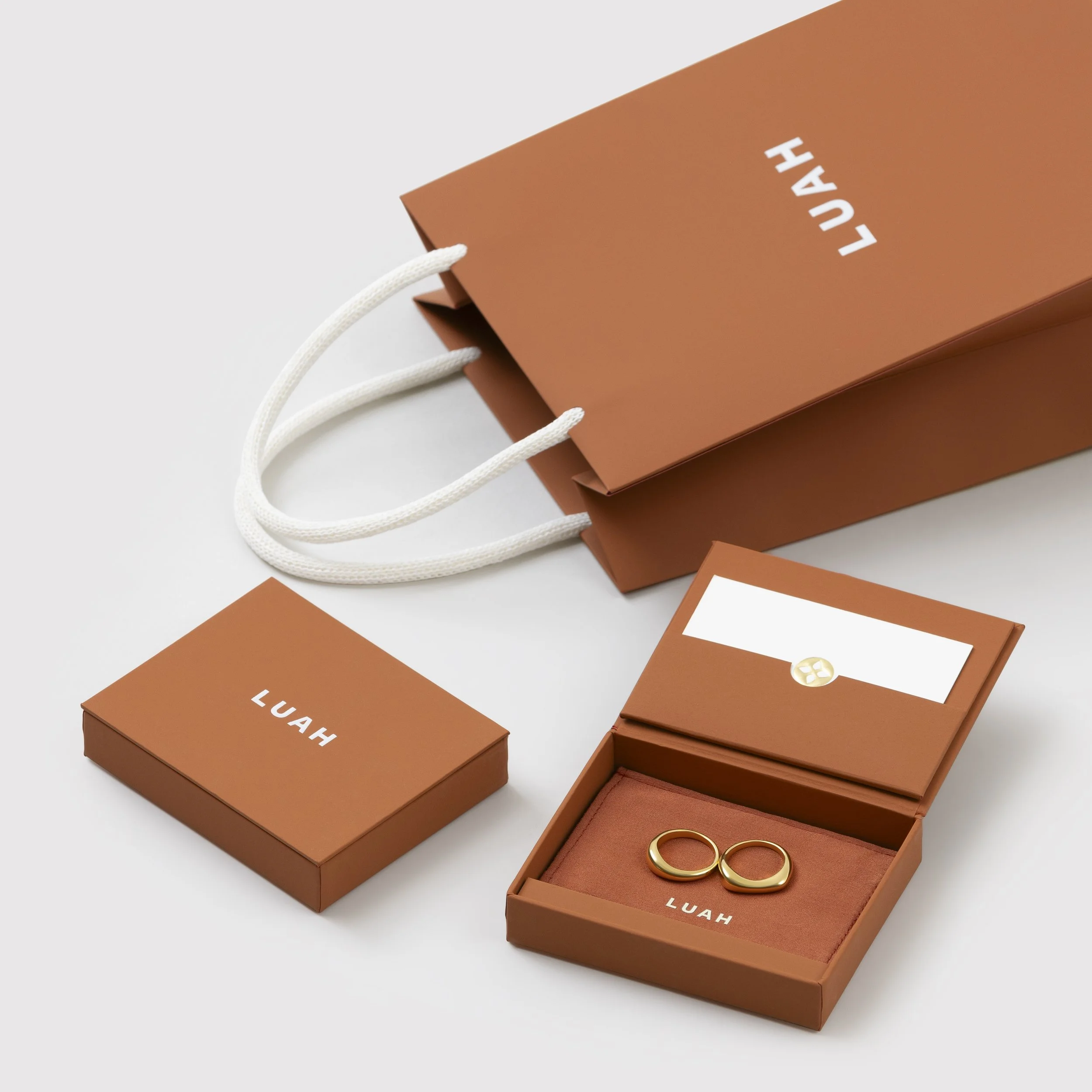 Gold rings in a brown jewelry box, with a matching brown shopping bag and a small brown box labeled 'LUAH' on a white surface.