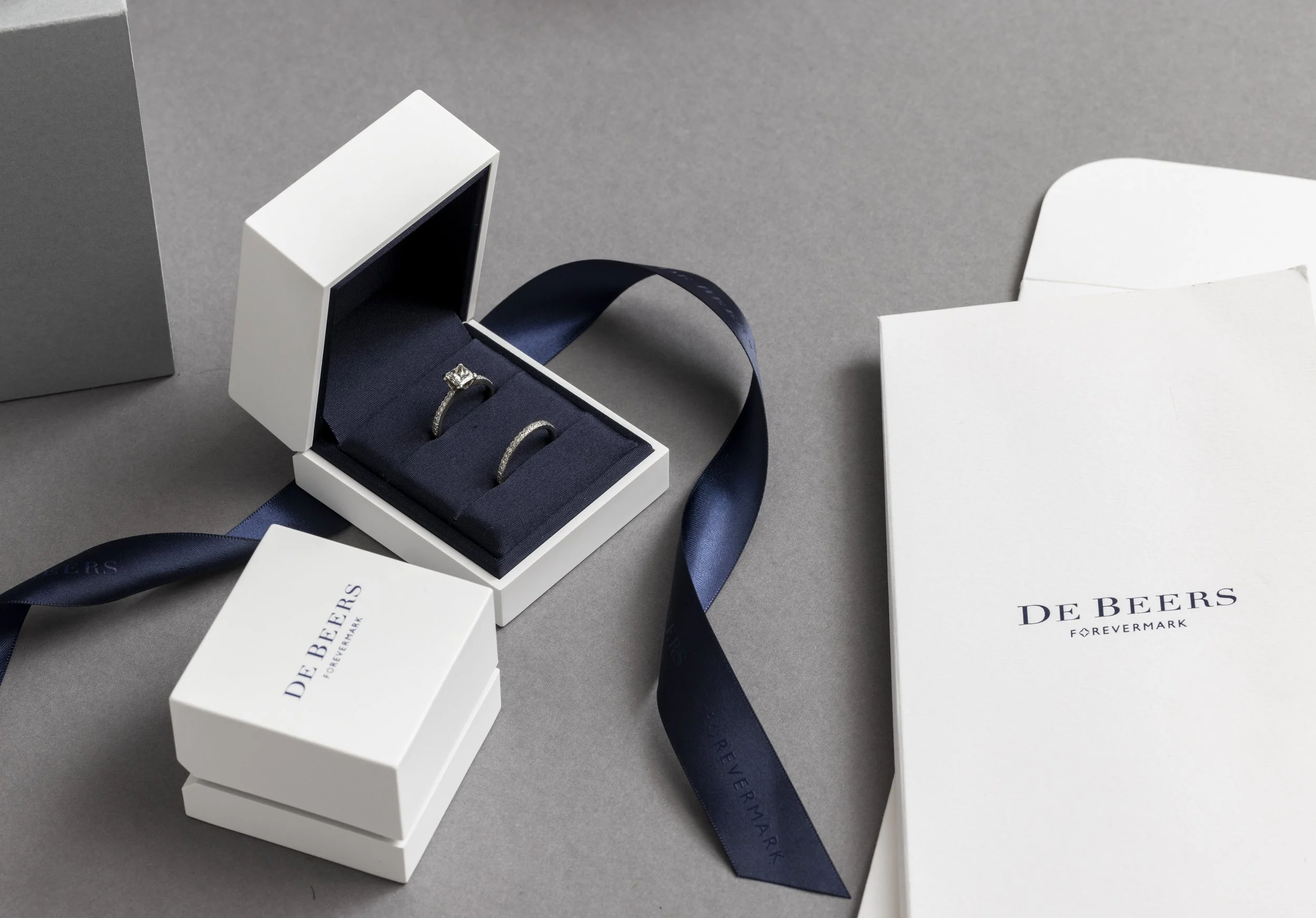 Open jewelry box with engagement and wedding rings, surrounded by branded packaging and a navy ribbon, on a gray surface.