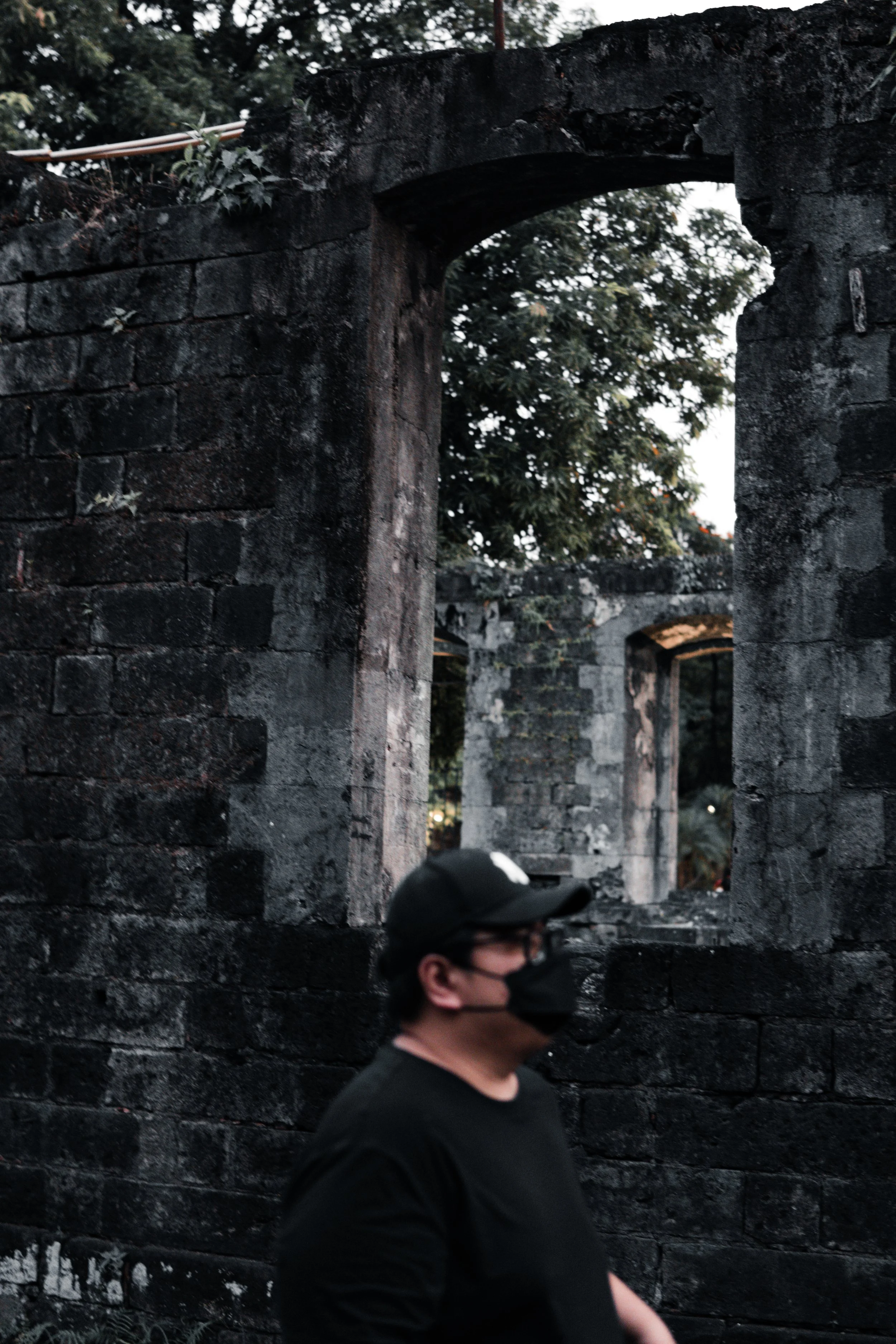 A person wearing a black face mask, black shirt, and black baseball cap walking past an old, weathered stone wall with arched windows and lush trees in the background.