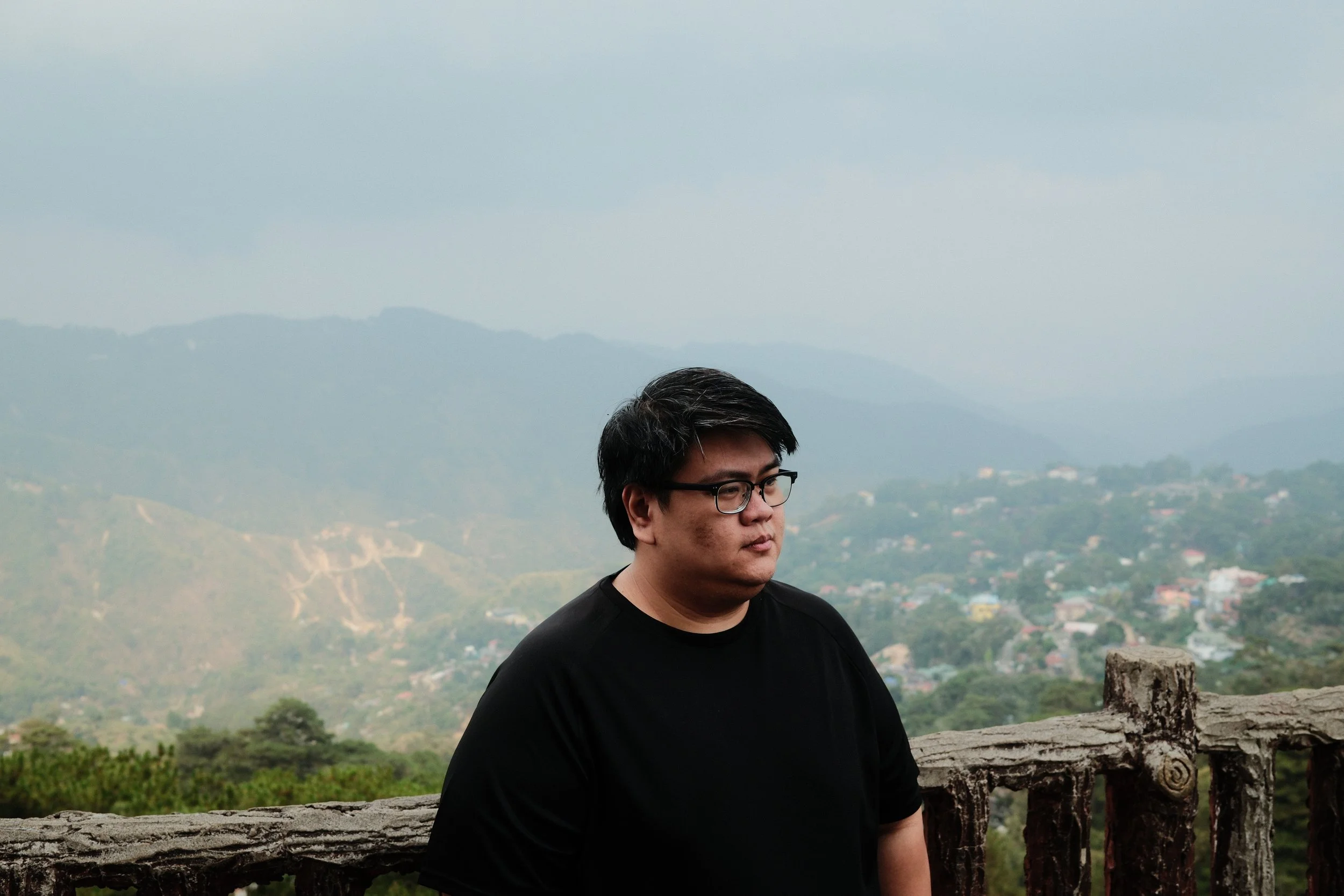 A man with glasses and short dark hair stands near a rustic wooden fence outdoors, overlooking a landscape of rolling hills and scattered houses with mountains in the distance under a cloudy sky.