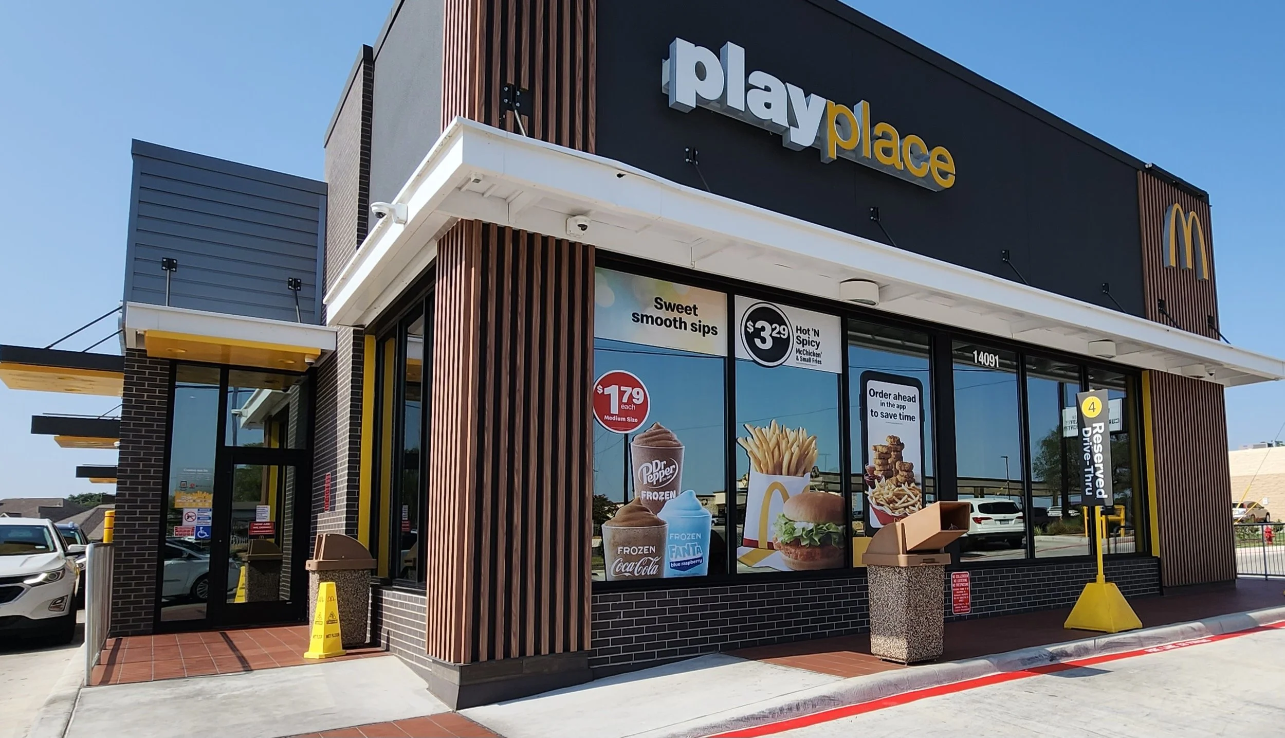 McDonald's restaurant exterior with signage, promotional posters for food items, and parking lot in the background under a clear blue sky.