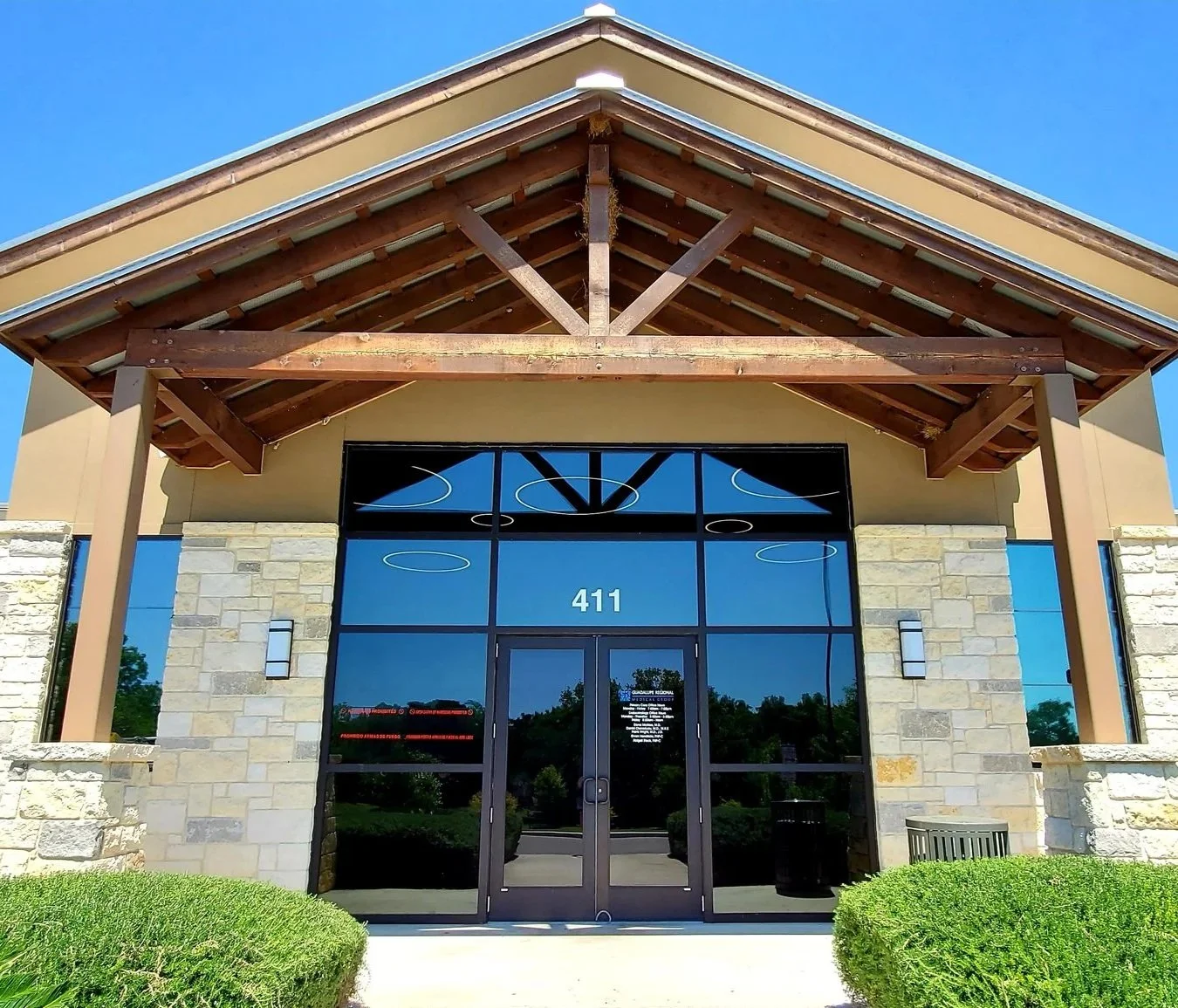 Front entrance of a modern building with a glass door, stone walls, and a wooden overhang, with the number 411 displayed above the door, surrounded by green shrubs.