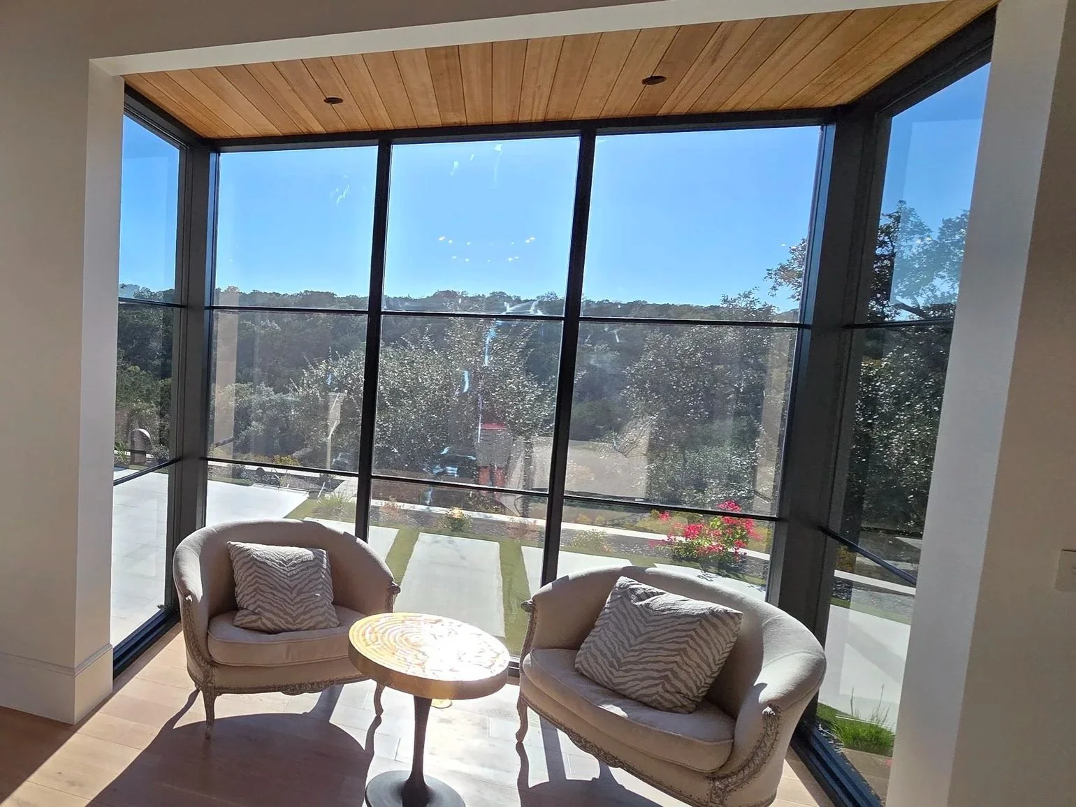 A cozy sitting area with two vintage-style armchairs and a small round wooden table in front of large floor-to-ceiling windows that offer a view of a sunny garden with trees and pink flowers, under a wooden ceiling.