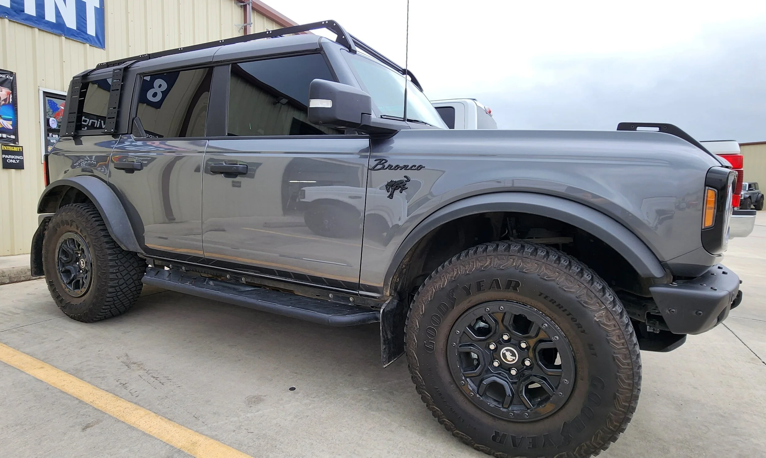 A gray Ford Bronco SUV parked outside a building with business signage. The vehicle has black wheels, large off-road tires, and black side steps.