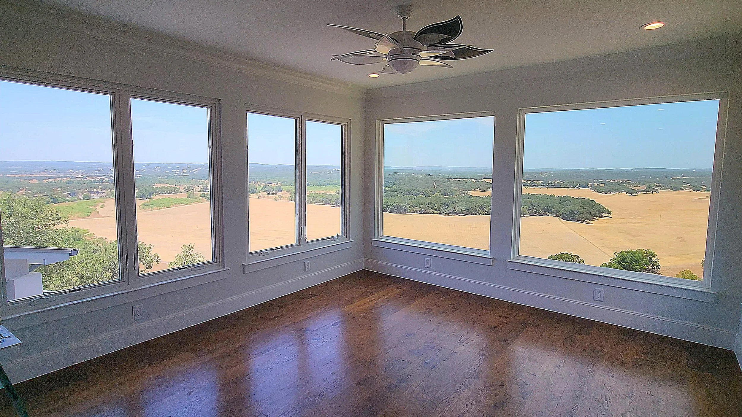 Empty room with large windows showcasing a scenic view of farmland and distant hills, hardwood floor, white walls, ceiling fan, and recessed lighting.