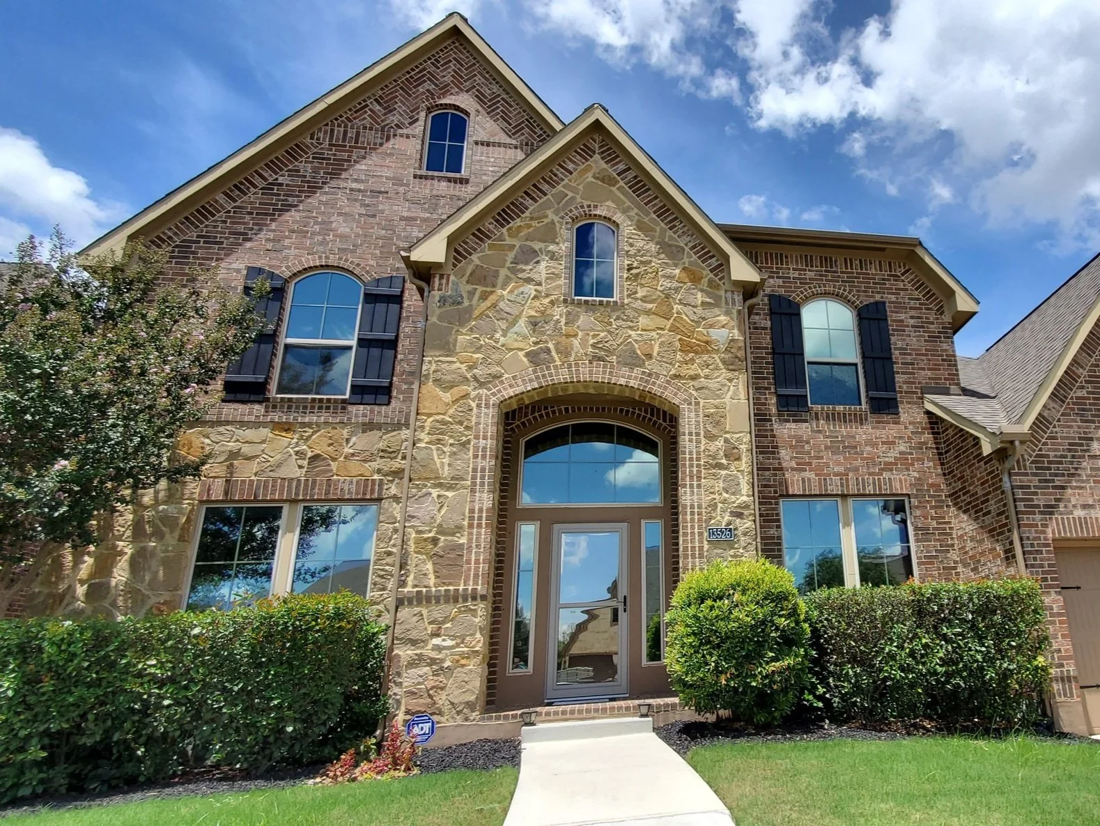 Front view of a two-story brick and stone house with a manicured lawn and bushes, under a blue sky with some clouds with solar control window tint to block heat from the sun from coming into the house. Shows reflective mirror window tint film by 3M