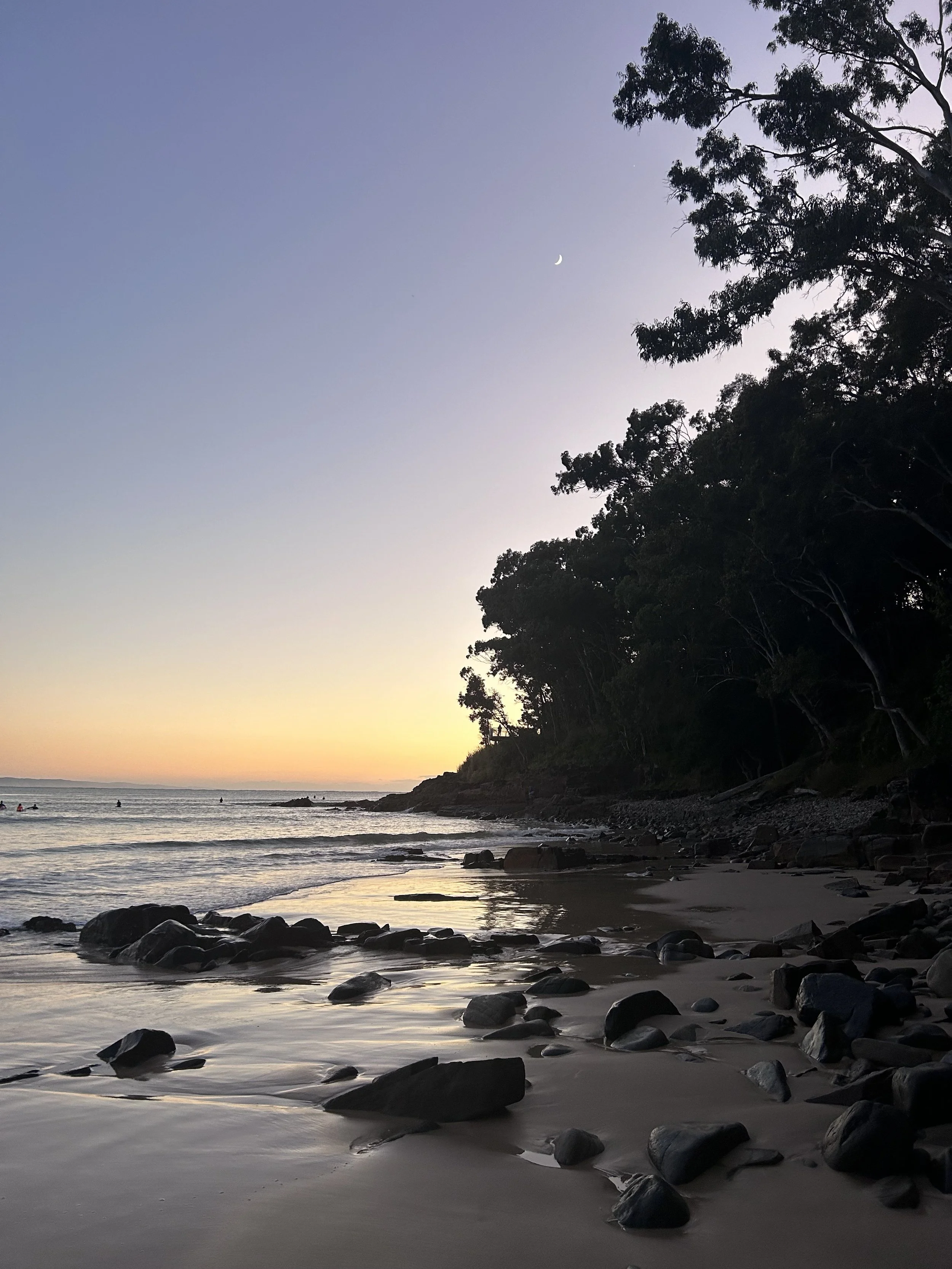 A beach scene at dusk with a view of the horizon, the sky transitioning from orange to blue, a crescent moon visible in the sky, rocks scattered along the shoreline, calm waves, and silhouettes of trees on the right side.