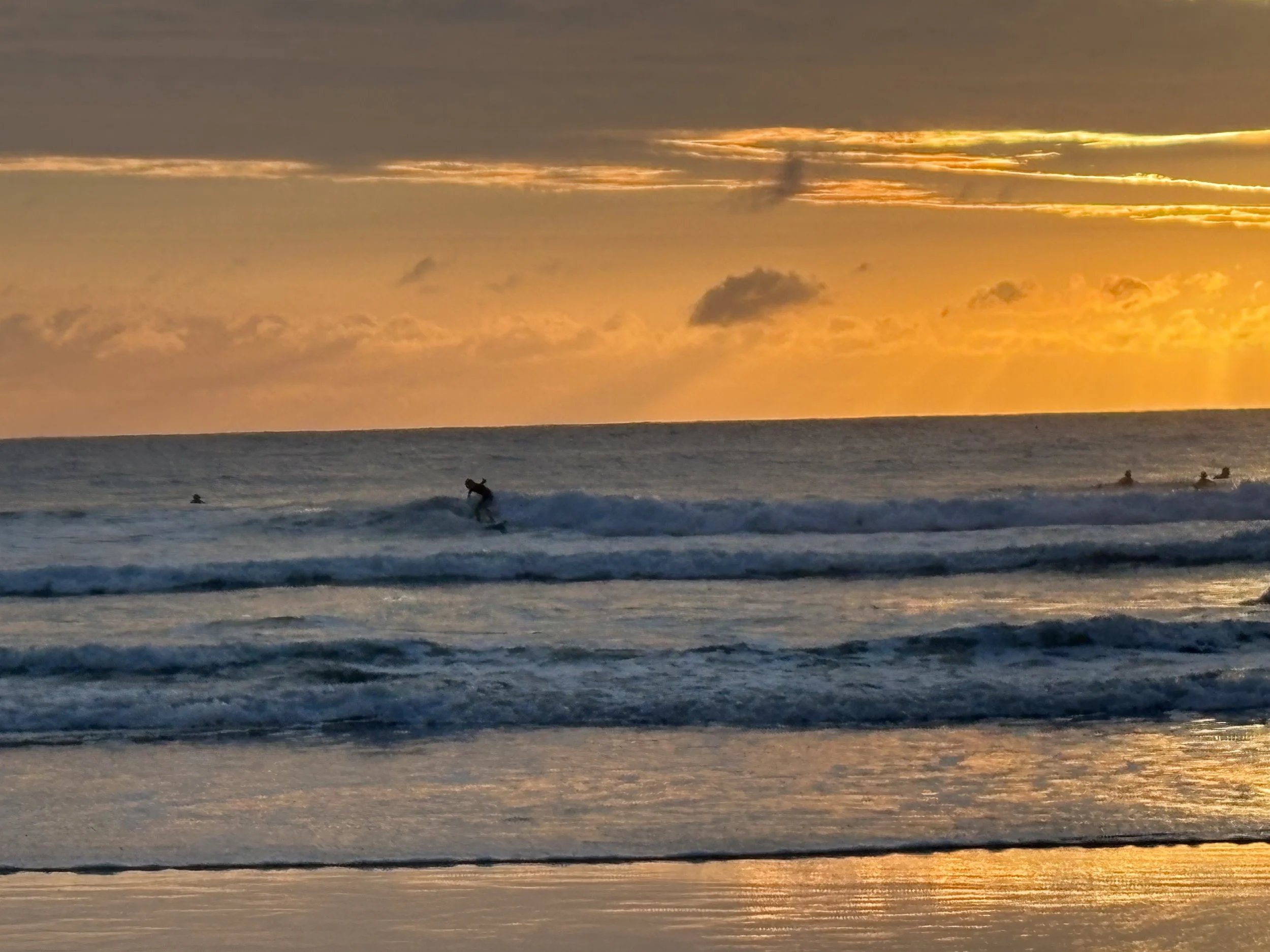 Sunset over the ocean with surfers riding the waves and others waiting, with orange and yellow hues in the sky.