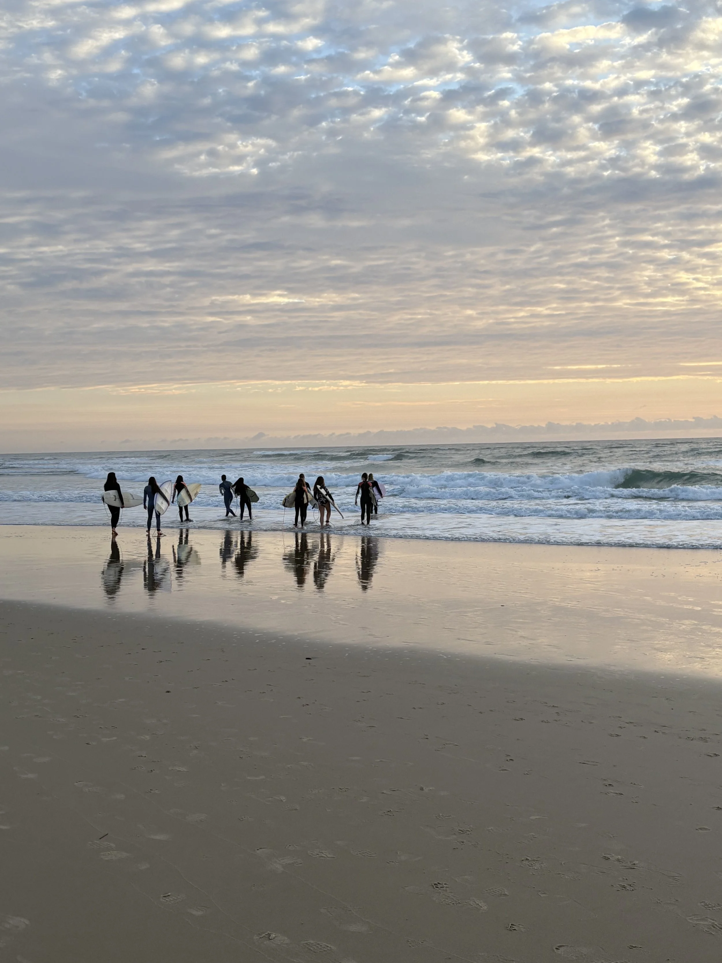A group of people with surfboards walking into the ocean at sunset on a beach, with cloudy sky and reflections on wet sand.