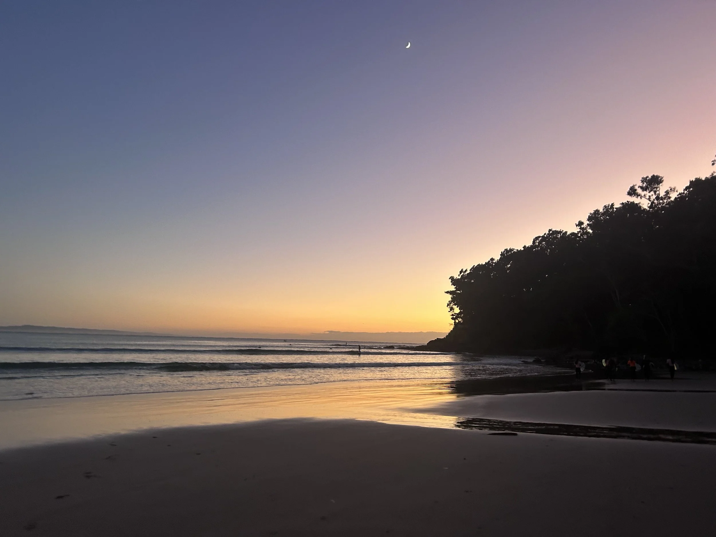 A beach at sunset with a clear horizon, calm waves, a small group of people on the shoreline, and a crescent moon in a colorful sky transitioning from sunset to twilight.