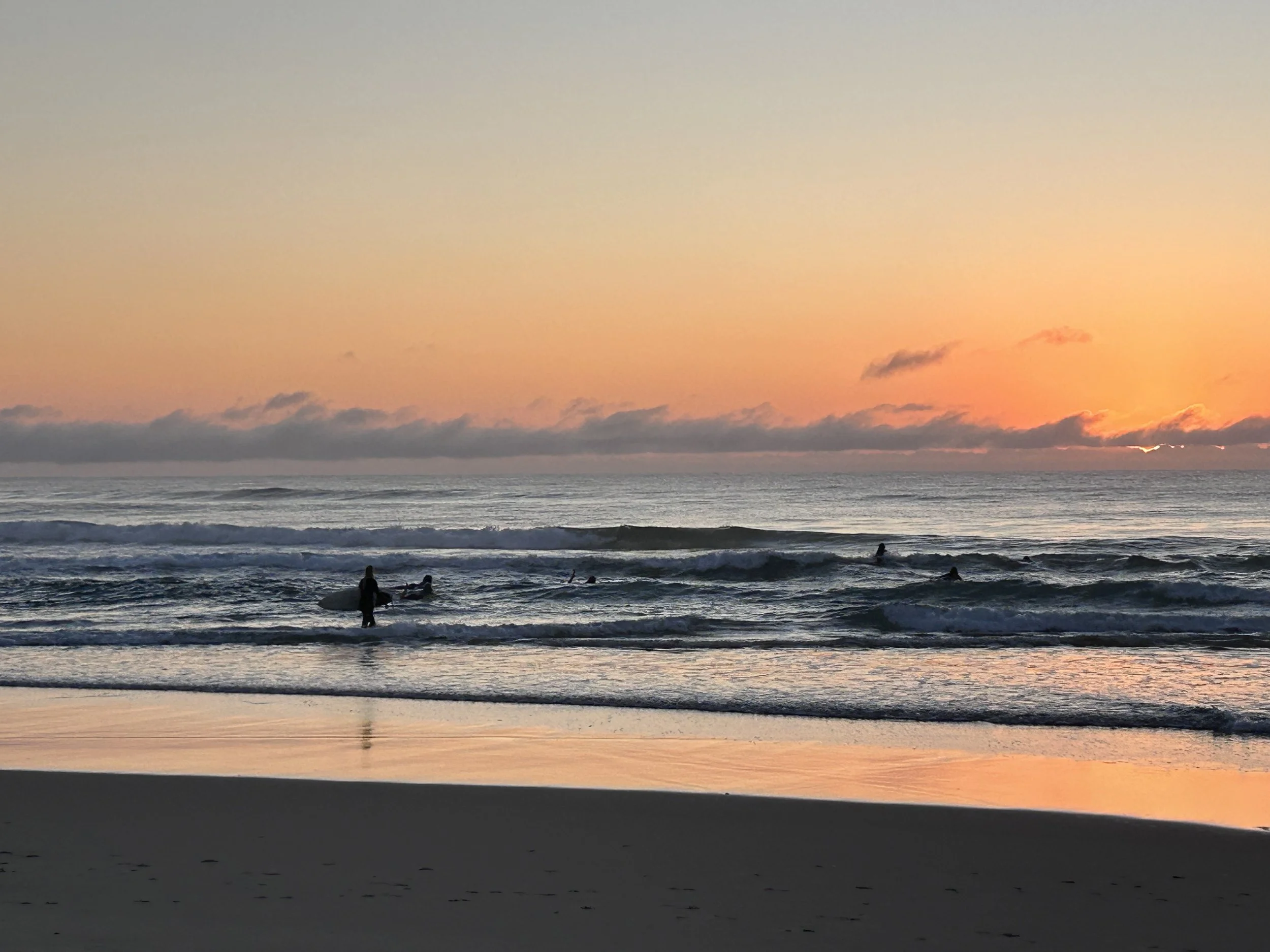 Sunset at the beach with a pink and orange sky, calm ocean waves, and silhouettes of surfers in the water.