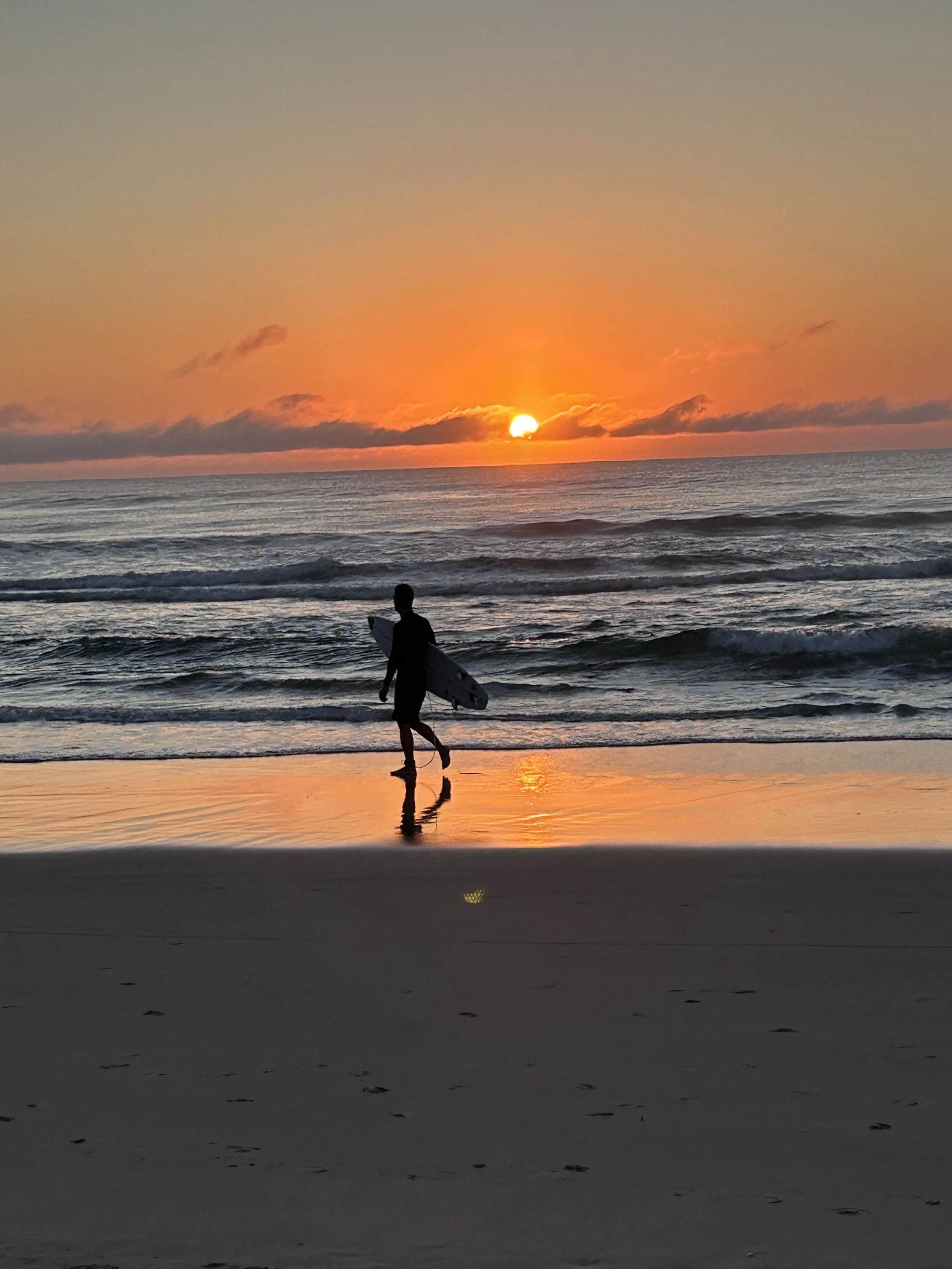 A person holding a surfboard walking along the beach at sunset, with the sun near the horizon and reflecting on the water.