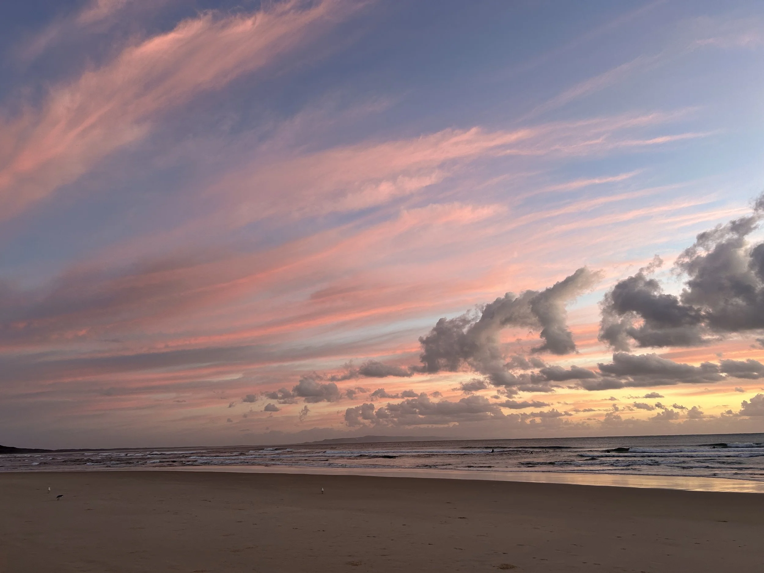 Sunset over a sandy beach with a partly cloudy sky in shades of pink, orange, and blue.