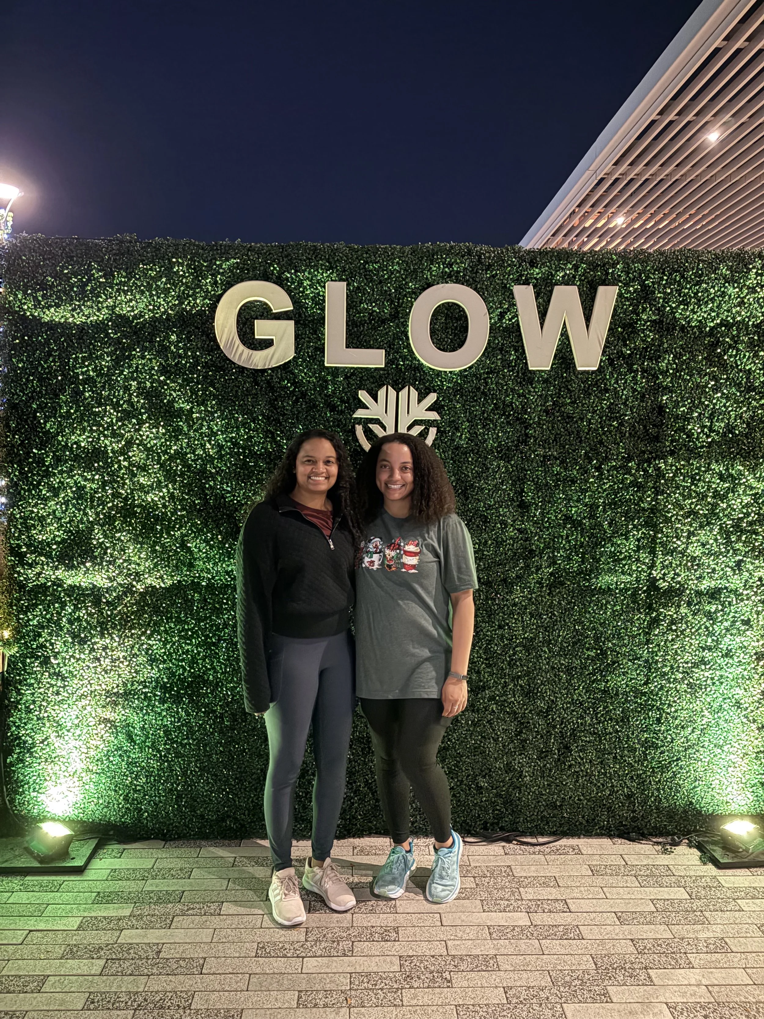 Lauren and Samantha standing in front of a giant GLOW sign in front of a makeshift hedge