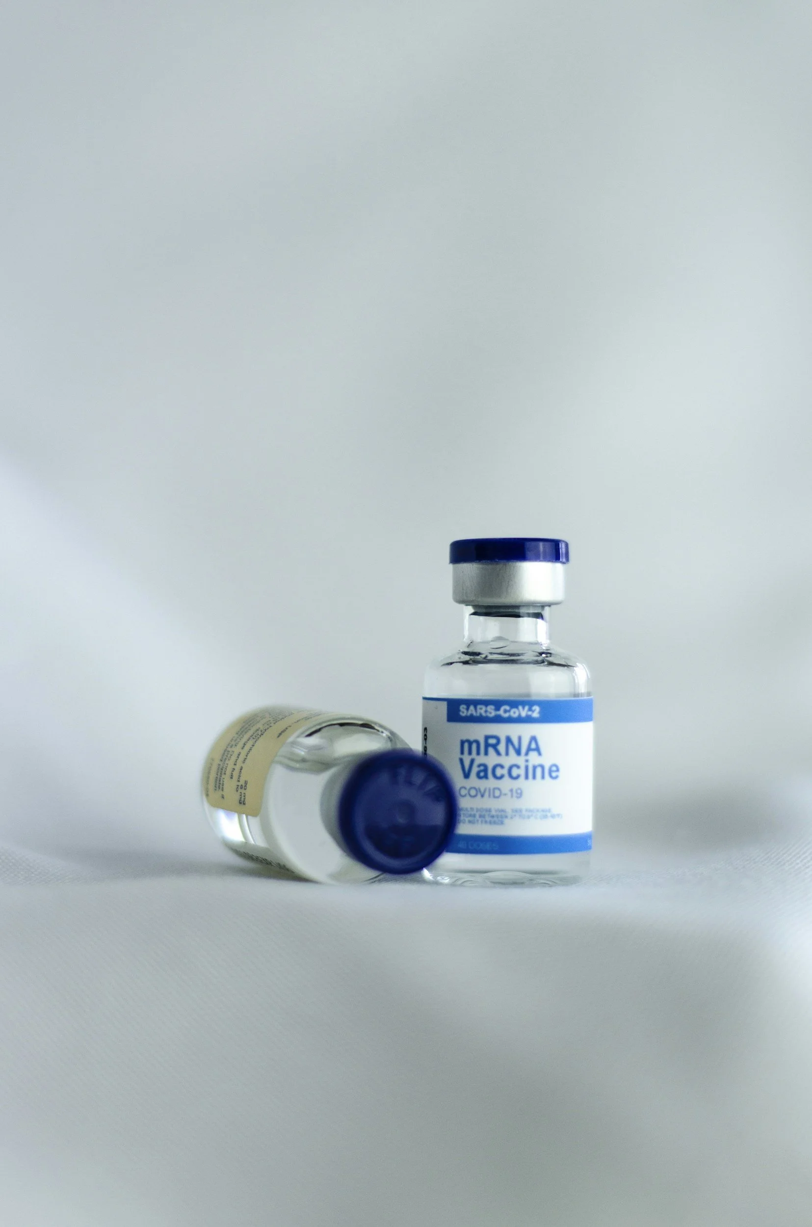Two vials of COVID-19 mRNA vaccine, one standing upright and the other lying on its side, with a plain background.