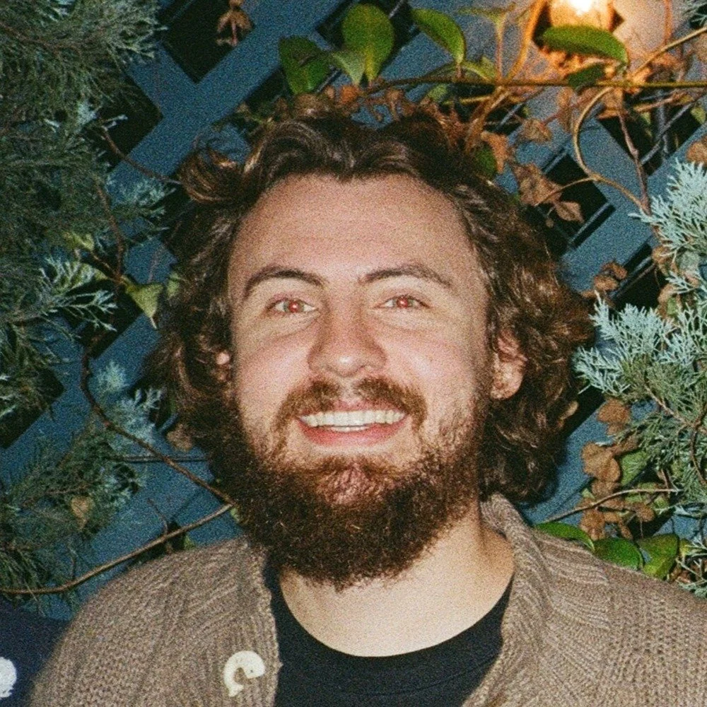 A smiling man with curly brown hair and a beard, wearing a brown jacket, standing in front of a background of green plants and a blue wooden fence.