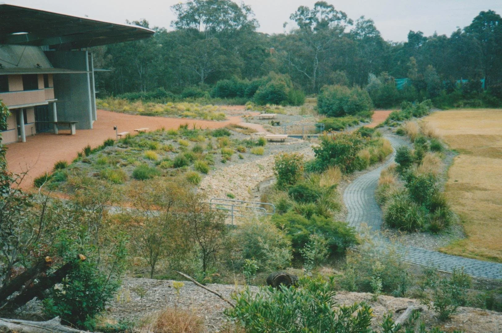 A landscaped garden area with a winding brick pathway, various bushes, and trees near a modern building with a curved roof.
