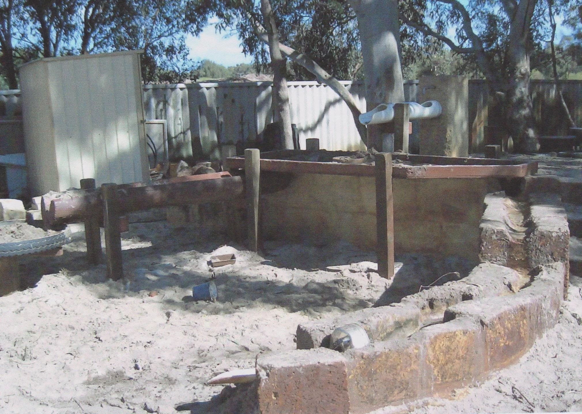 A makeshift outdoor fire pit with a brick border, wooden logs, and a metal pipe, surrounded by sand and trees, with a metal fence and some trees in the background.