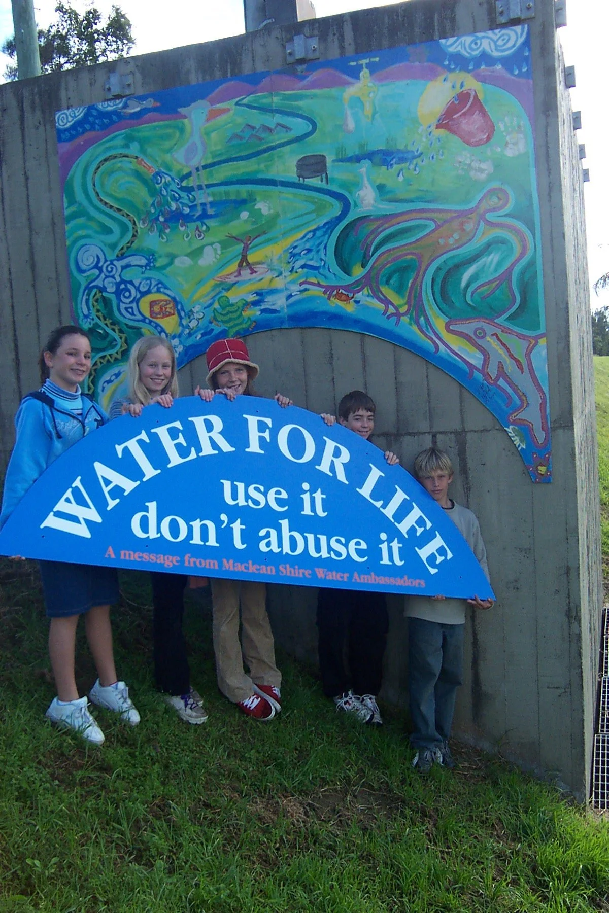 Group of children holding a blue banner with the message 'Water for Life, use it don't abuse it,' in front of a colorful mural depicting water-related scenes and animals.
