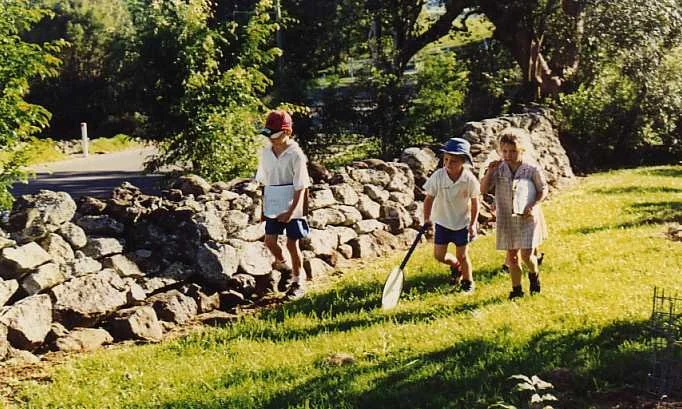 Three children dressed in school uniforms, carrying school supplies, walking on a grassy area near a stone wall in a sunny outdoor setting.