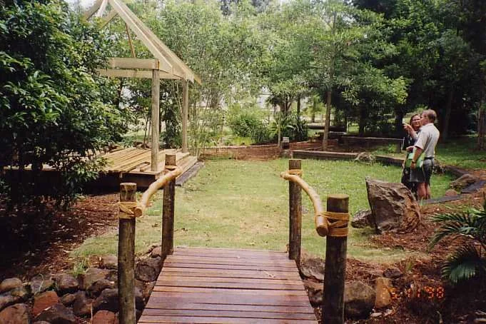 A small wooden bridge with rope railing in a park or garden, with trees and two people standing near a large rock on the right side.