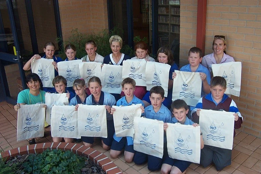 Group of school children and teachers holding reusable cloth bags with no water symbols, outside a school building.