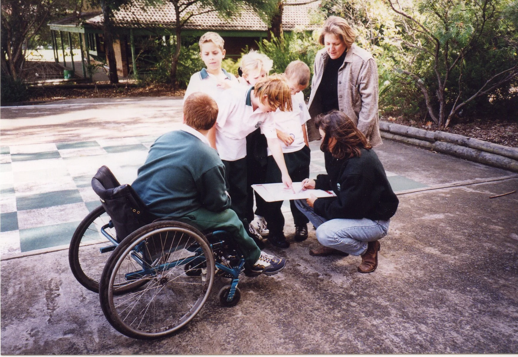A group of children and adults gathered outdoors around a clipboard, with one child in a wheelchair. They are engaged in a discussion or activity, outside on a paved area with trees and a building in the background.