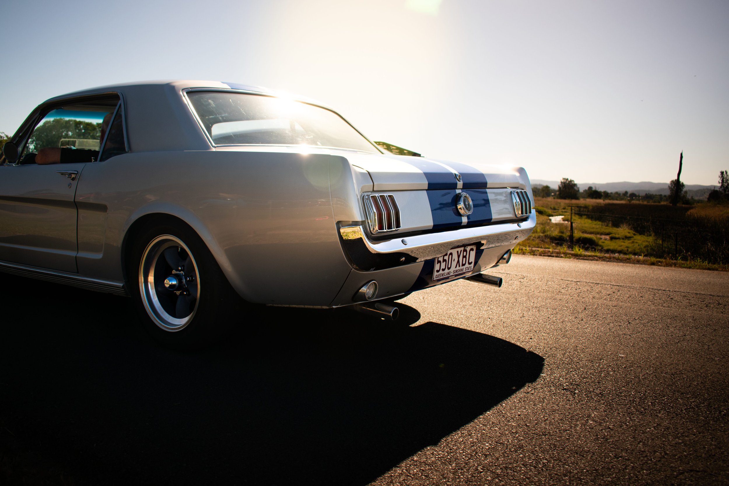 Classic 1966 Ford Mustang coupe with a distinctive blue racing stripe, parked on a sunlit road, emphasizing its vintage automotive design and style