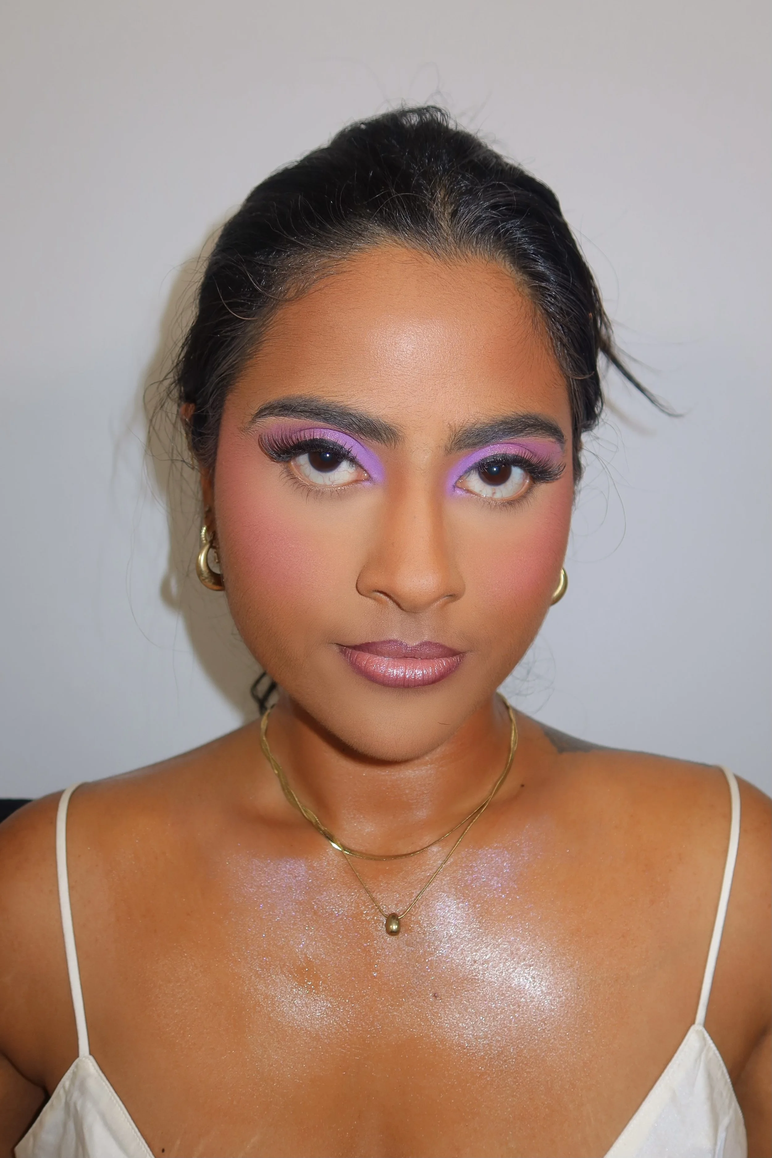 A woman with dark hair styled back, wearing makeup with purple eyeshadow, pink blush, and glossy lips, dressed in a white top with thin straps, accessorized with gold jewelry, and having a natural expression against a plain background.