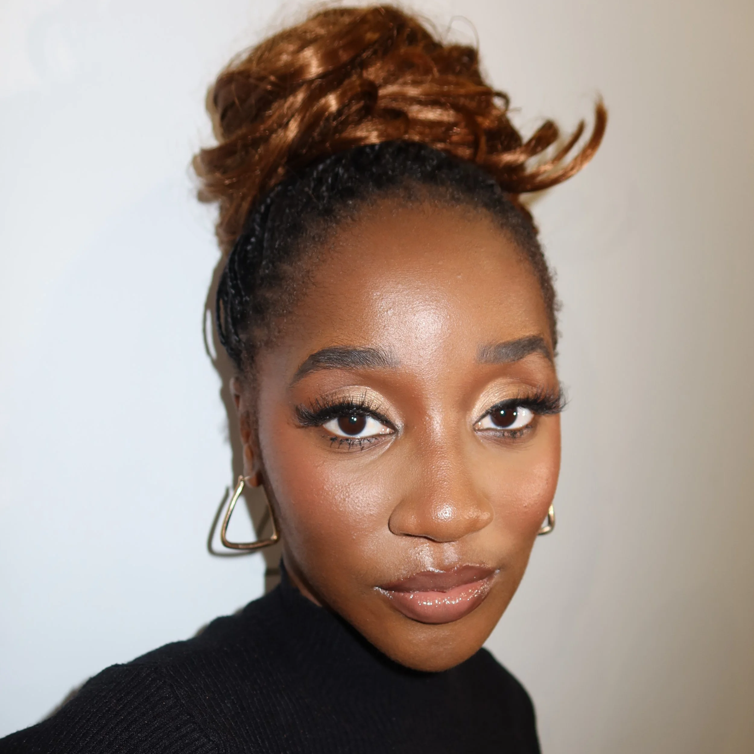 Close-up portrait of a young African American woman with styled brown hair in an updo, wearing makeup and gold hoop earrings, against a plain background.
