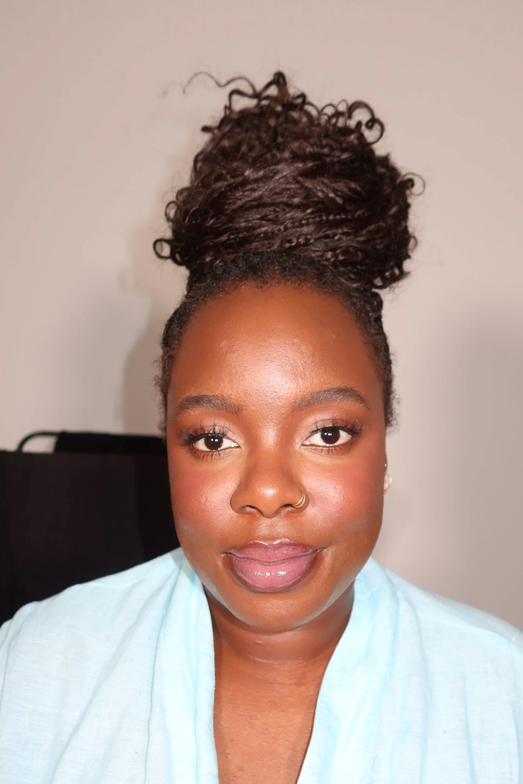 A woman with dark brown skin, styled with long, twisted hair gathered into a high bun, wearing a light blue top and small earrings, looking directly at the camera.