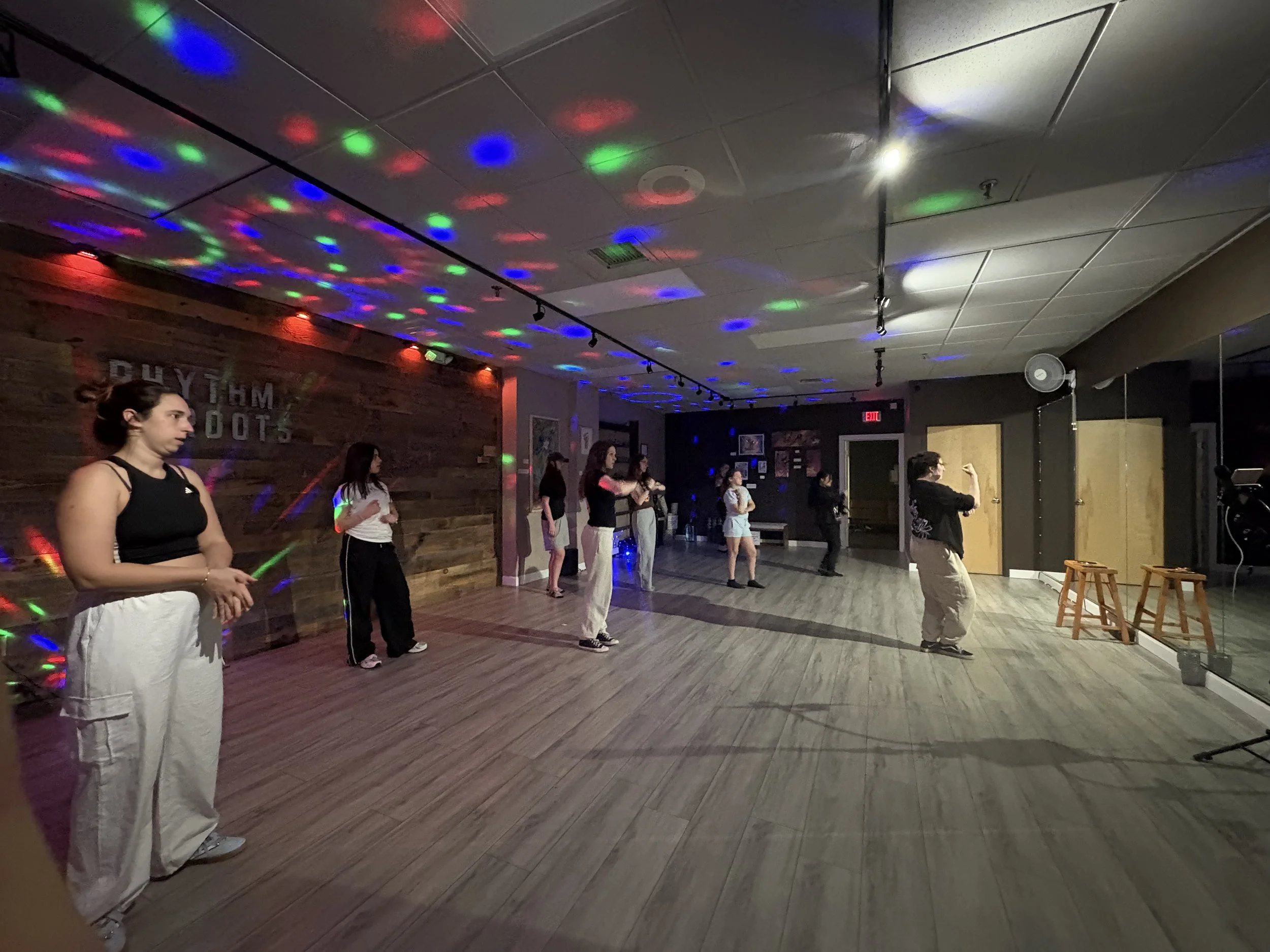 Group of women practicing dance moves in a studio with colorful lights and a wooden wall.