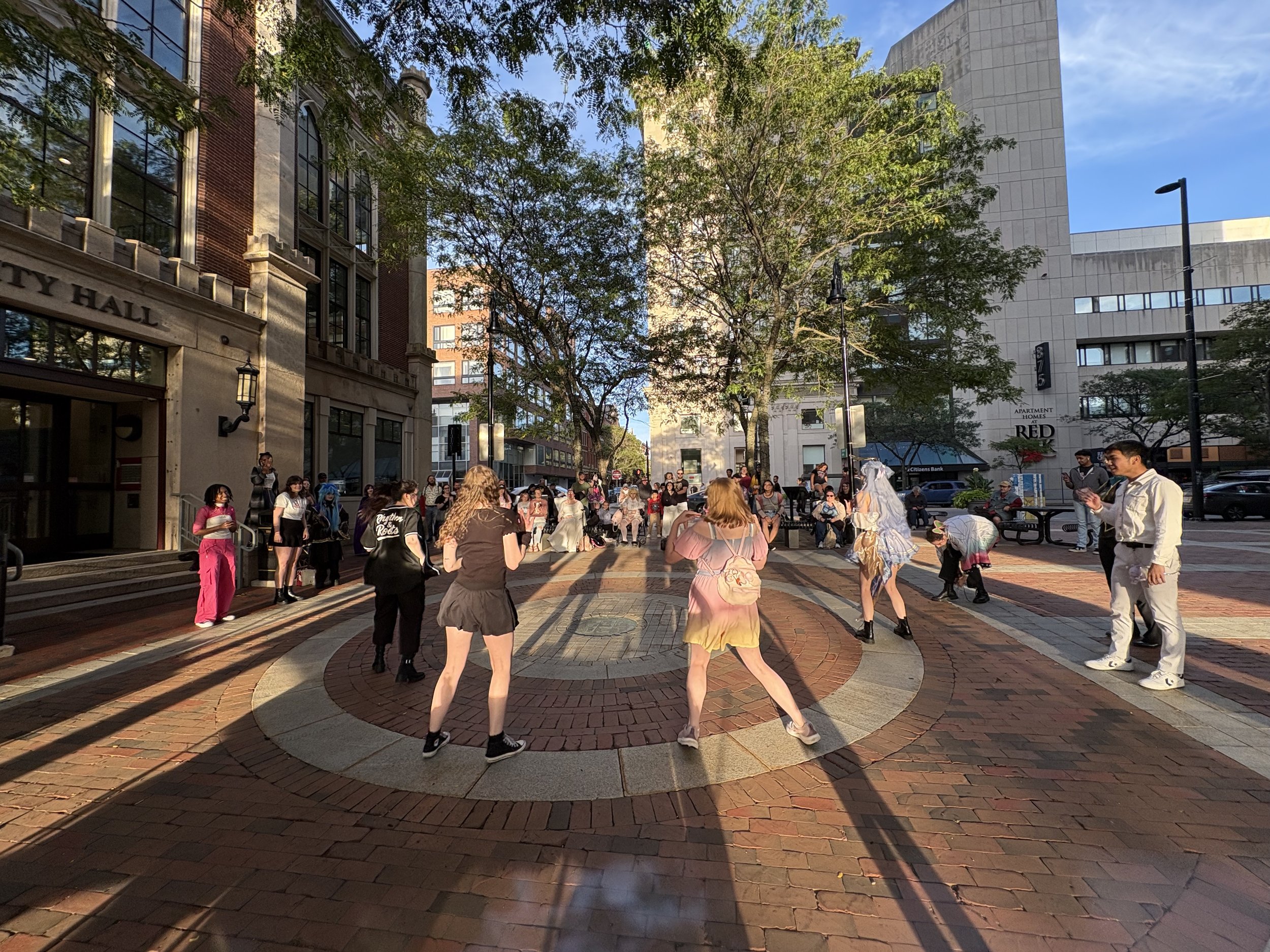 People dancing in a public square during sunset, with onlookers watching, surrounded by buildings and trees.
