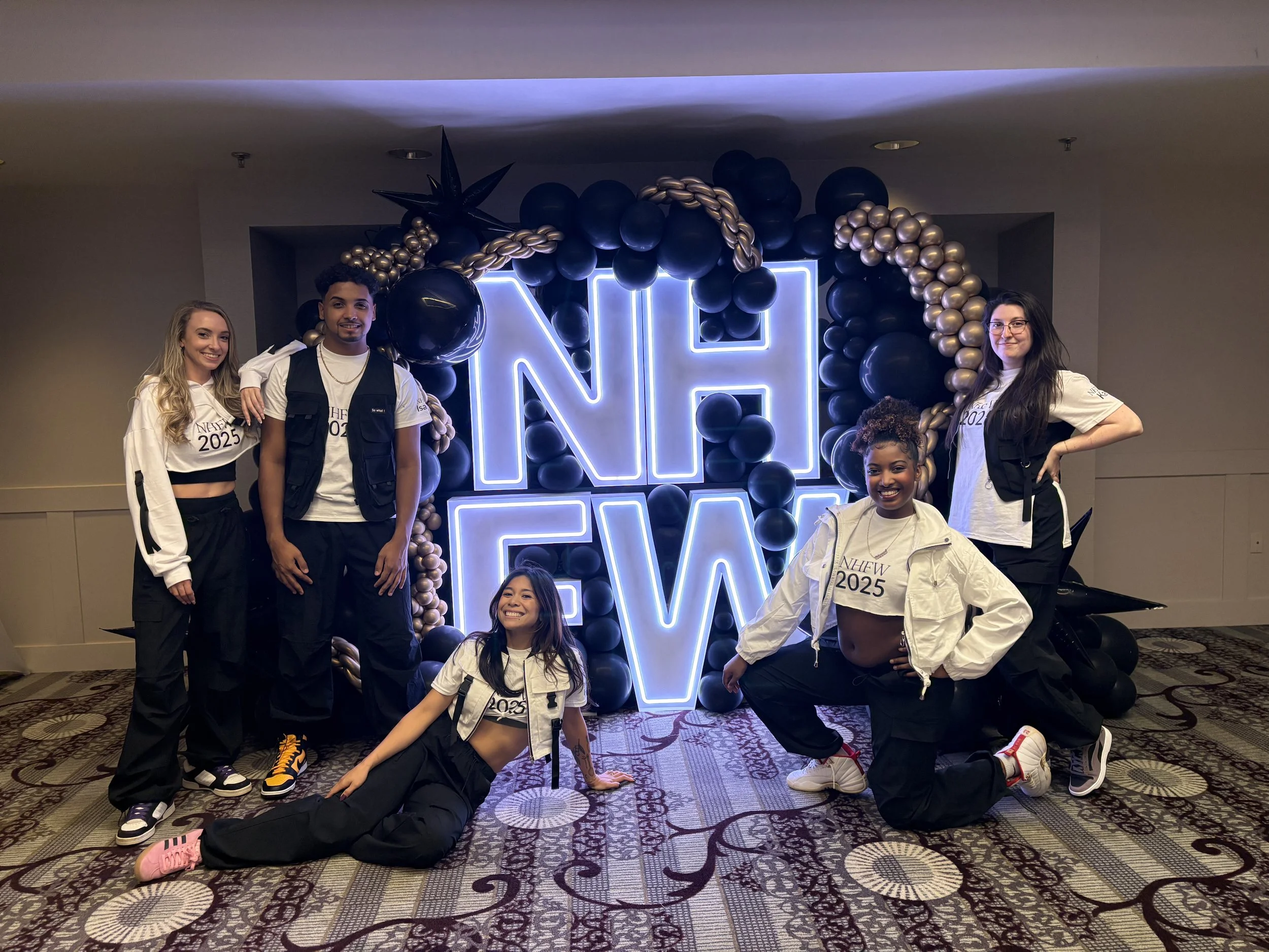 Group of six young people posing in front of a large illuminated neon sign that reads 'NHEW 2025' with black and gold balloon decorations.