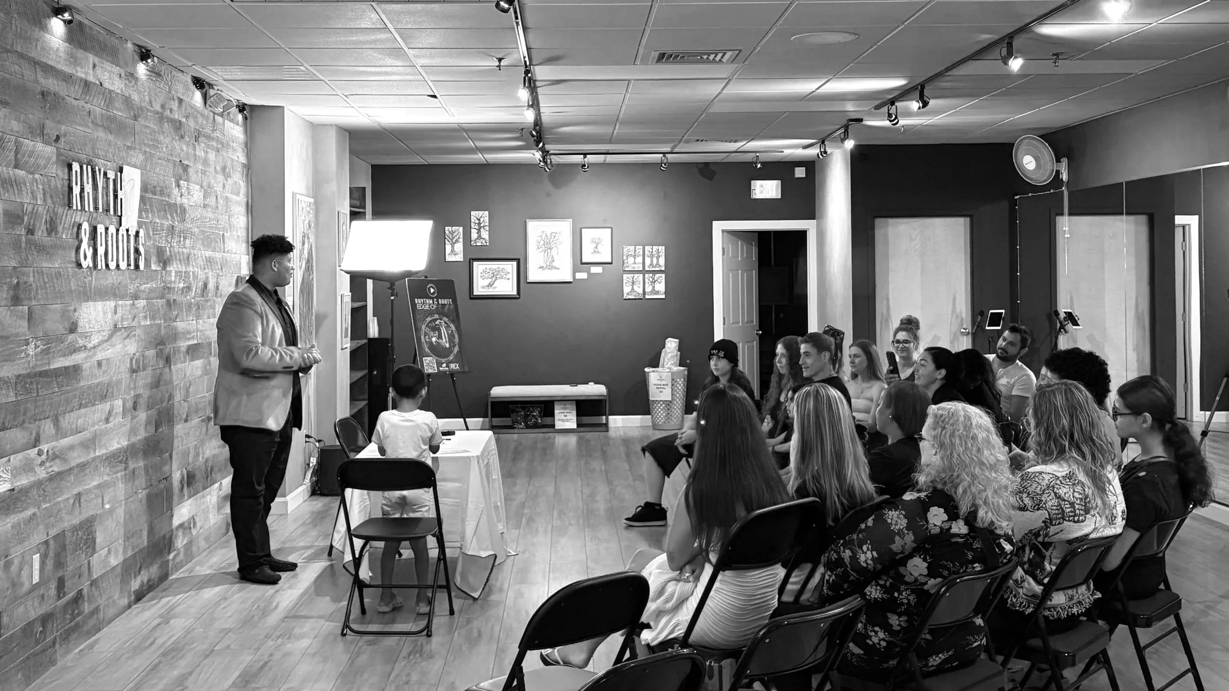 A man in a blazer speaking to a group of people seated in a room with wooden walls and framed pictures on the wall. A child is seated at a small table near the speaker.