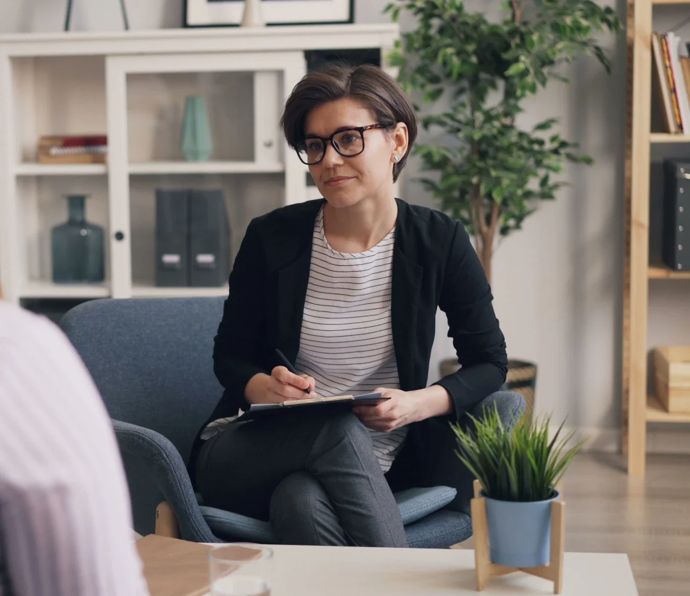 A female therapist with glasses writing on a notepad while listening to a client during a counseling session.