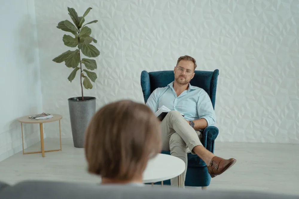 Male therapist sitting in a blue armchair holding a notepad during a counseling session with a client.