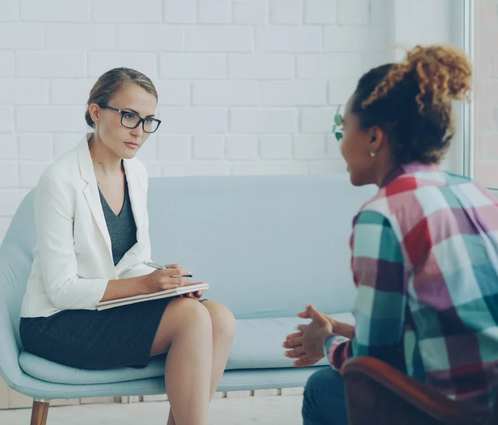 Female therapist in glasses taking notes on a notepad while listening to a client during a counseling session.