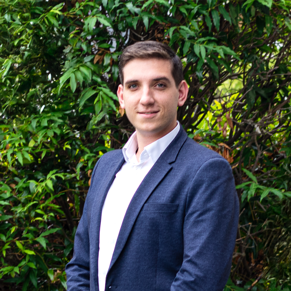 A young man wearing a navy suit jacket and white shirt standing outdoors in front of green leafy bushes.