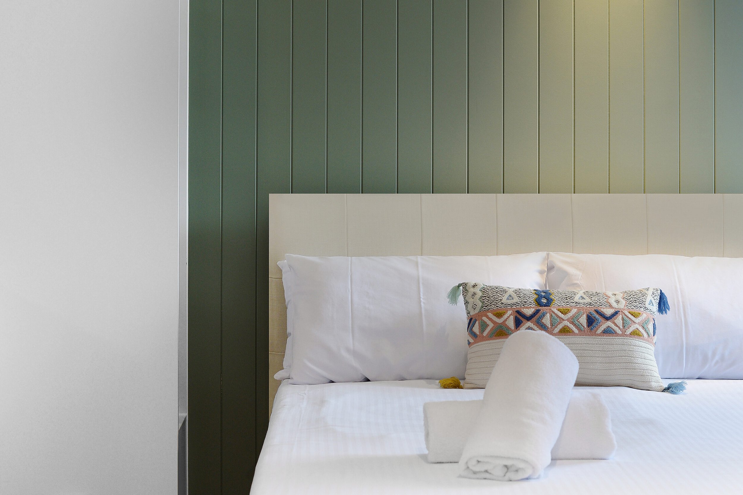 A neatly made bed with white pillows, a decorative pillow with colorful geometric embroidery, and rolled white towels on top of the bed, in front of a green paneled wall.