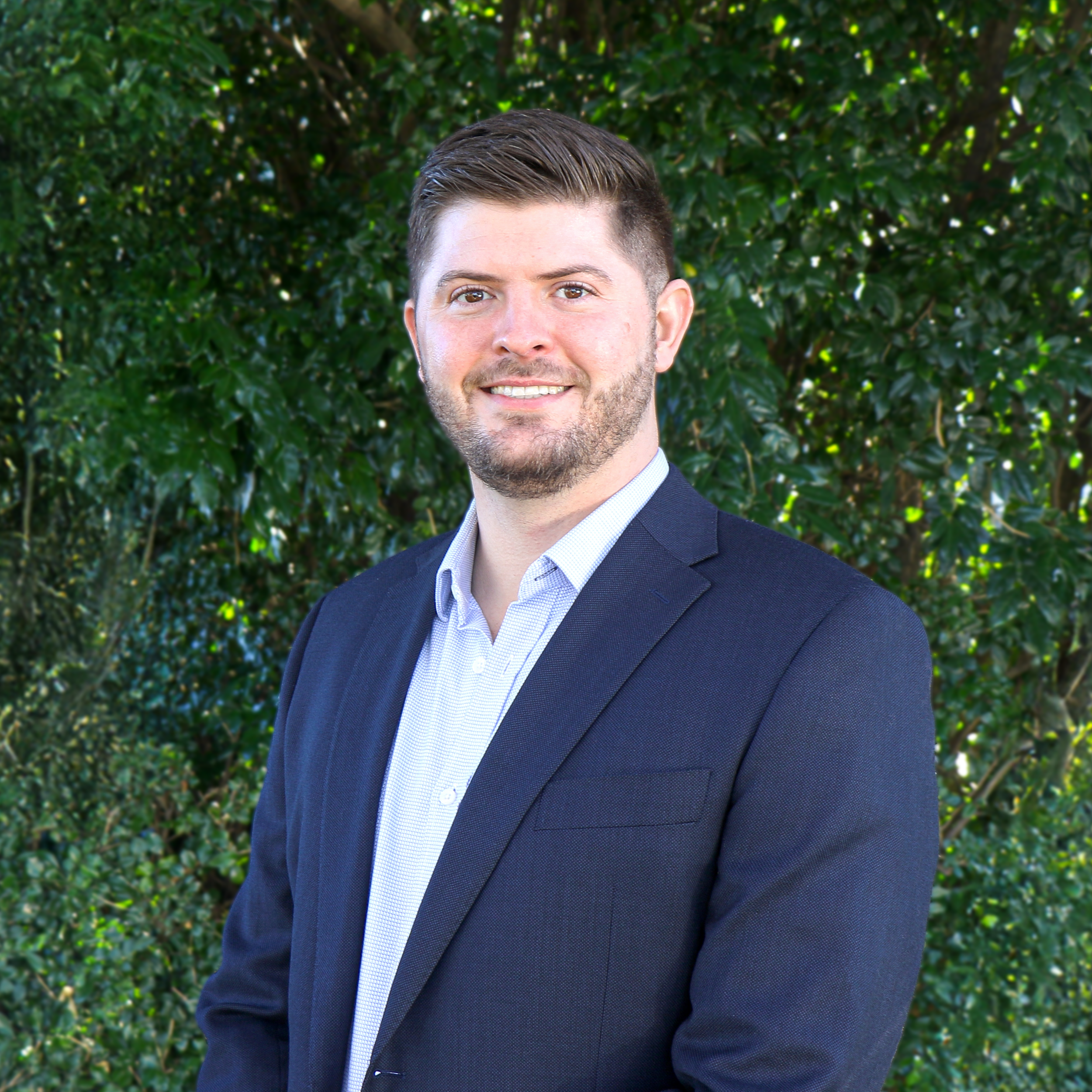 Professional man with brown hair and beard wearing a dark suit jacket and light blue dress shirt, standing outdoors in front of green foliage, smiling.
