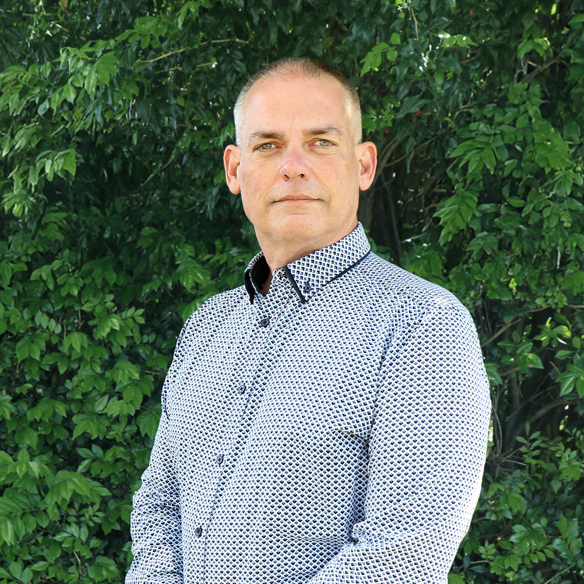 A man with short hair and a serious expression standing outdoors in front of green leafy bushes, wearing a patterned button-up shirt.