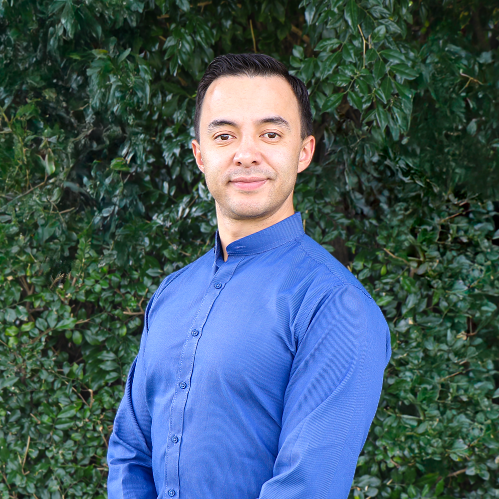 Portrait of a man in a blue button-up shirt standing outdoors with green leafy bushes in the background.